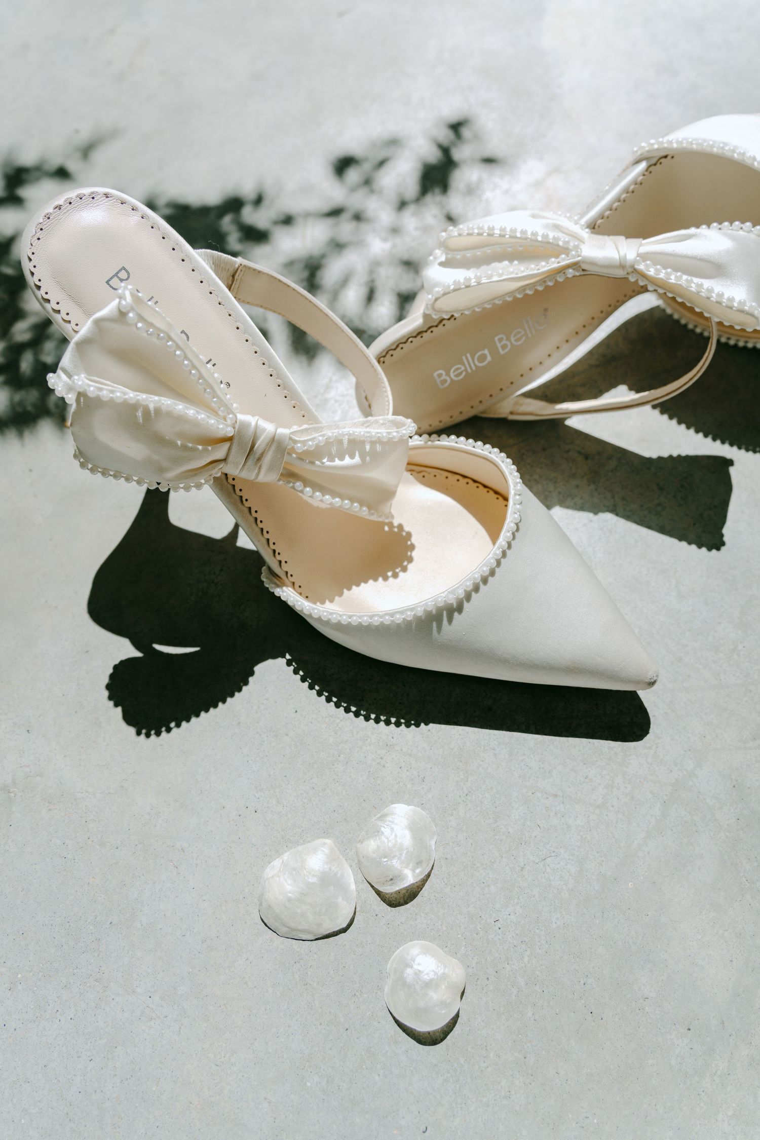 Bella Belle white bridal heels with pearl bow detail on marble surface with crystal earrings.