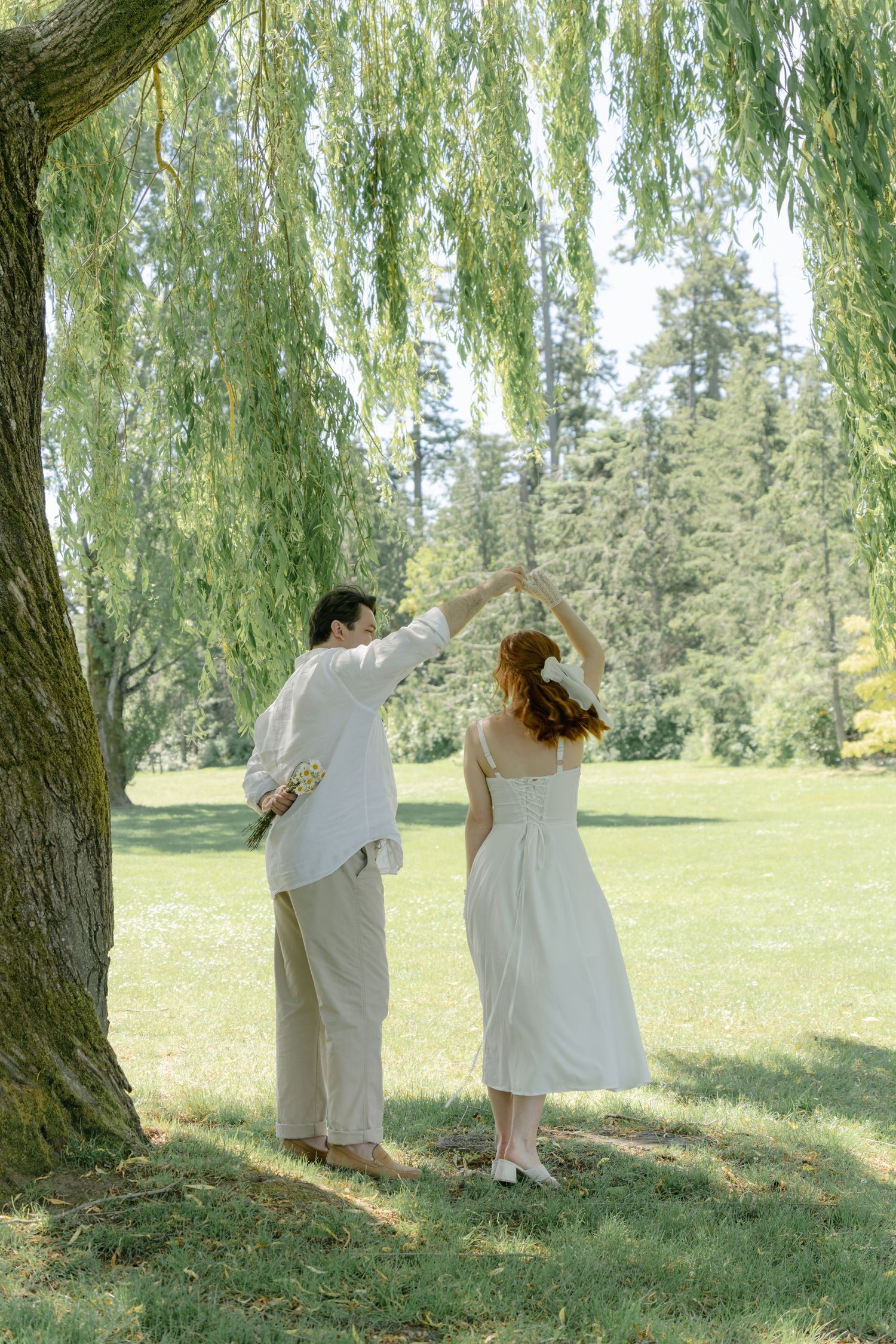 A couple in light clothing dances together under a weeping willow tree in a peaceful garden setting.