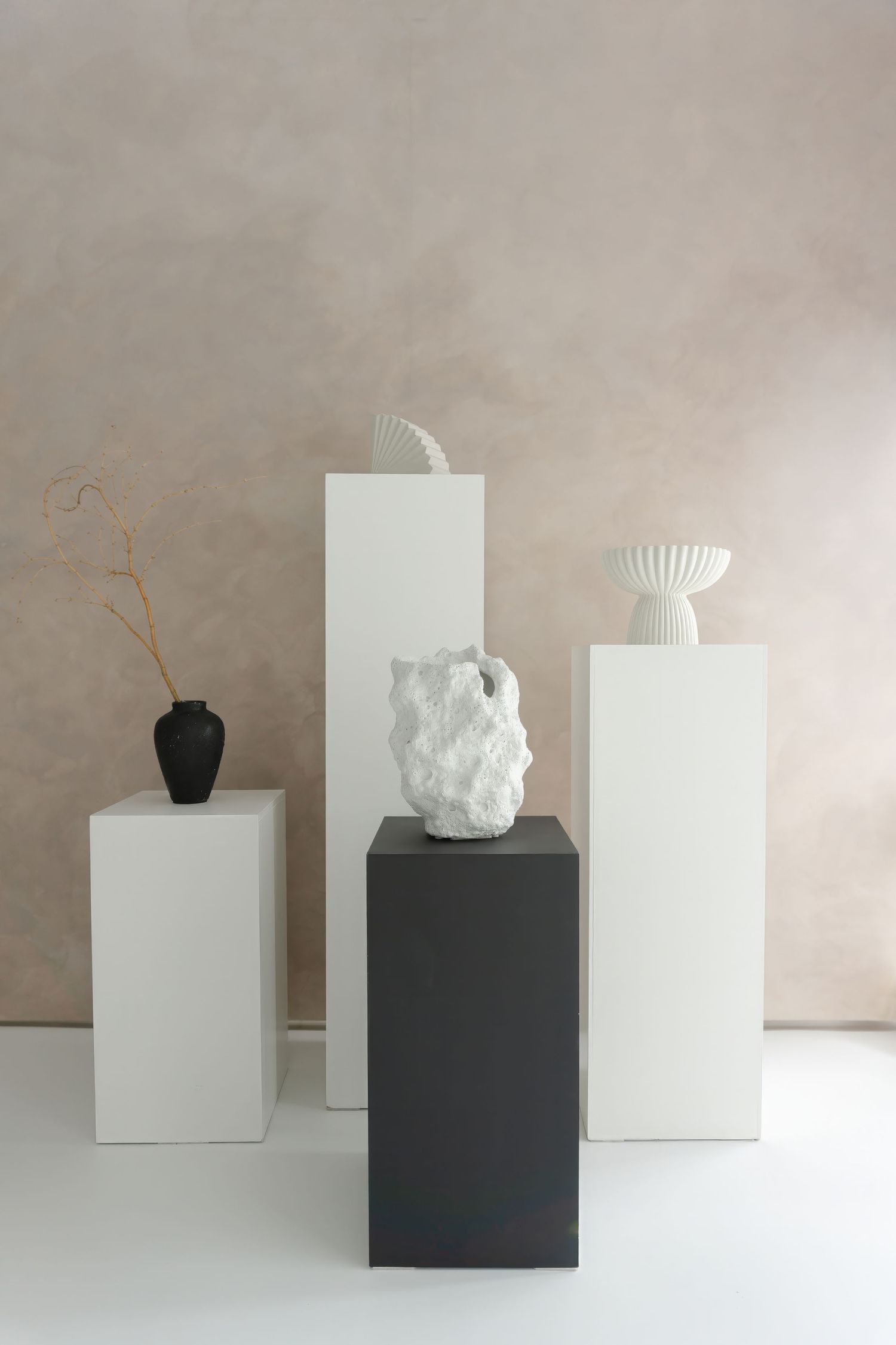 Modern decorative vases and sculptures displayed on white and black pedestals against neutral wall.