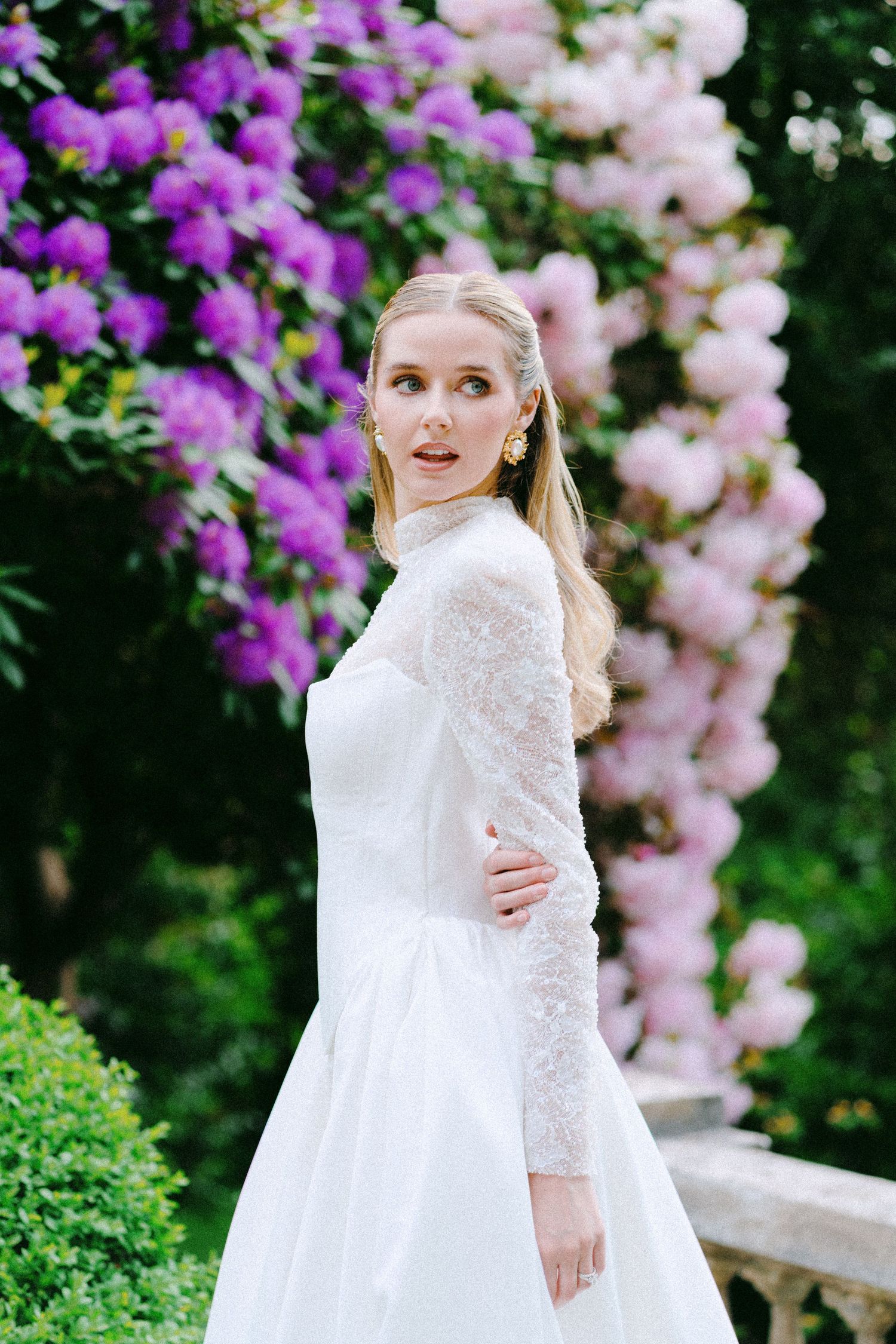 Blonde bride in white lace long-sleeve gown poses before vibrant purple and pink flowering trees in a garden.