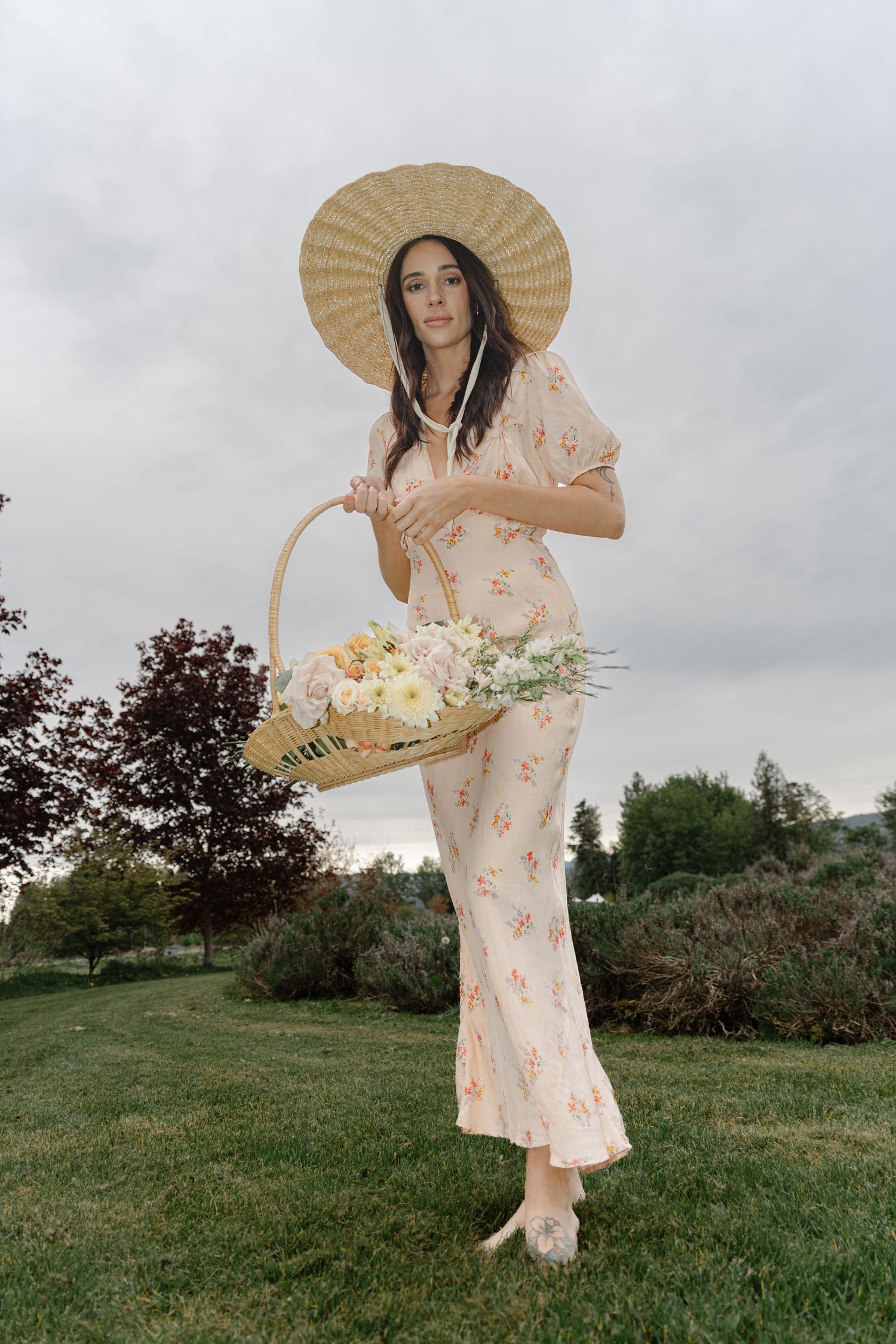 Woman in peach floral dress poses with straw hat in green field in Vancouver Island