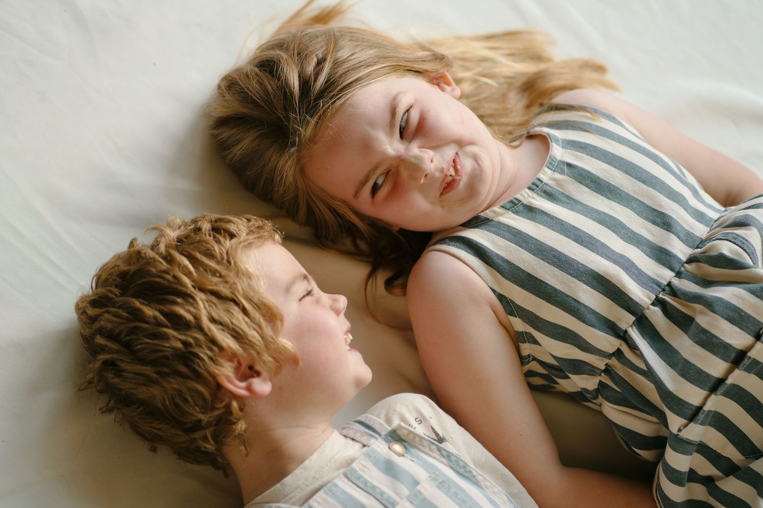 Two children lying on a bed smiling at each other, a curly-haired boy and a girl in a striped top.