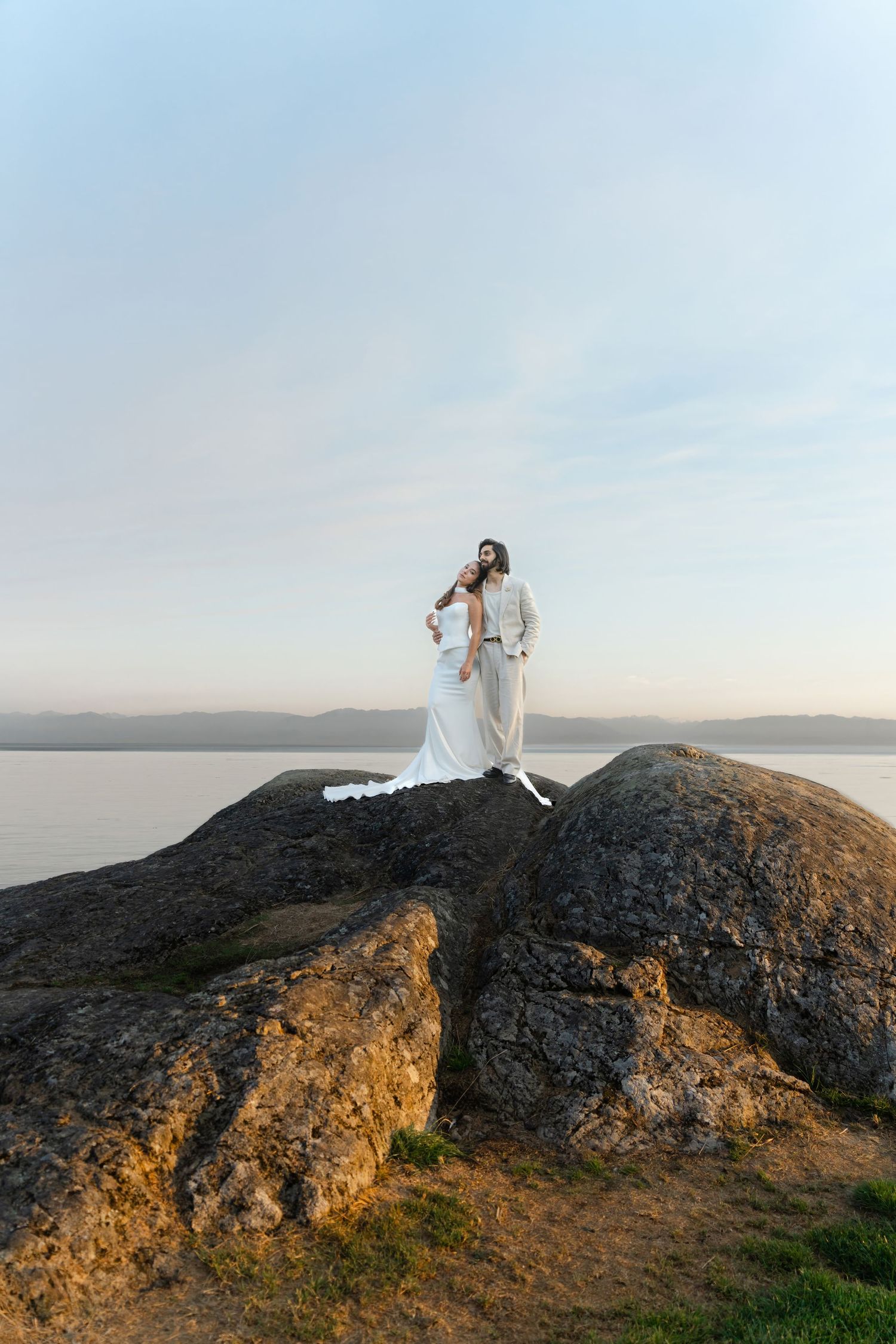 A couple stands atop a scenic coastal cliff at sunset, creating a romantic silhouette against the ocean backdrop.