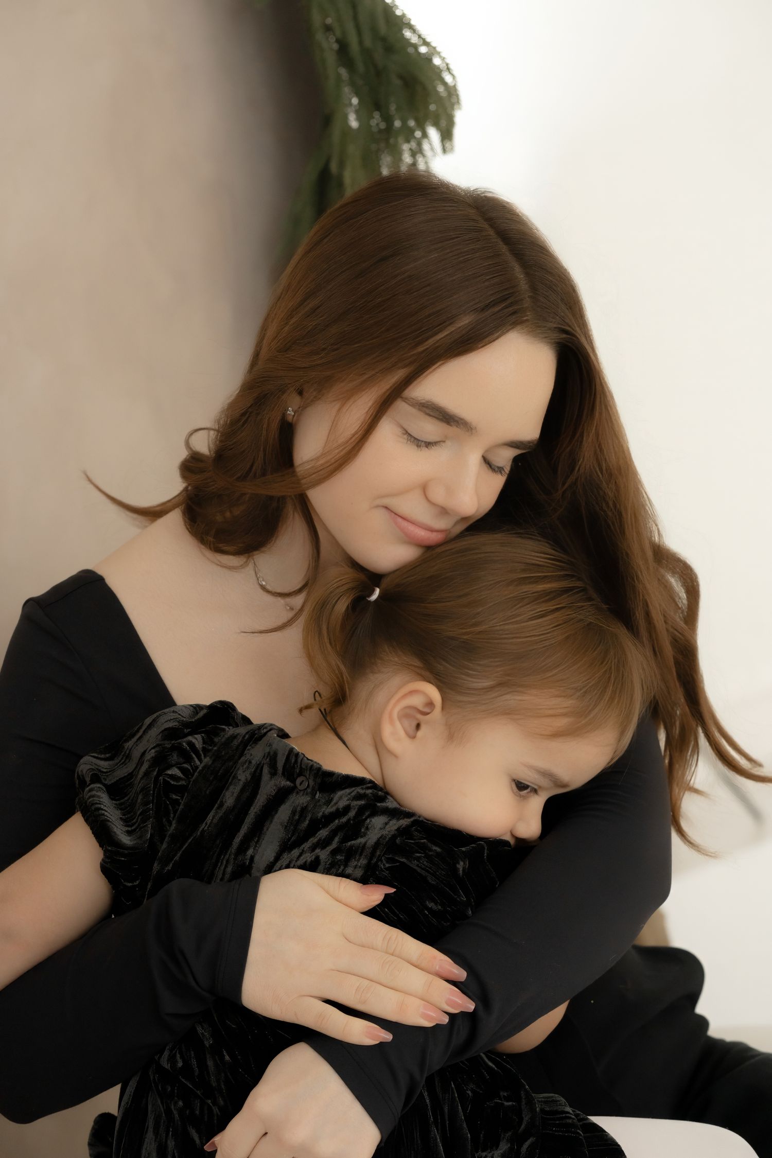 A young mother in black dress tenderly embraces her toddler daughter, both wearing matching dark velvet outfits.