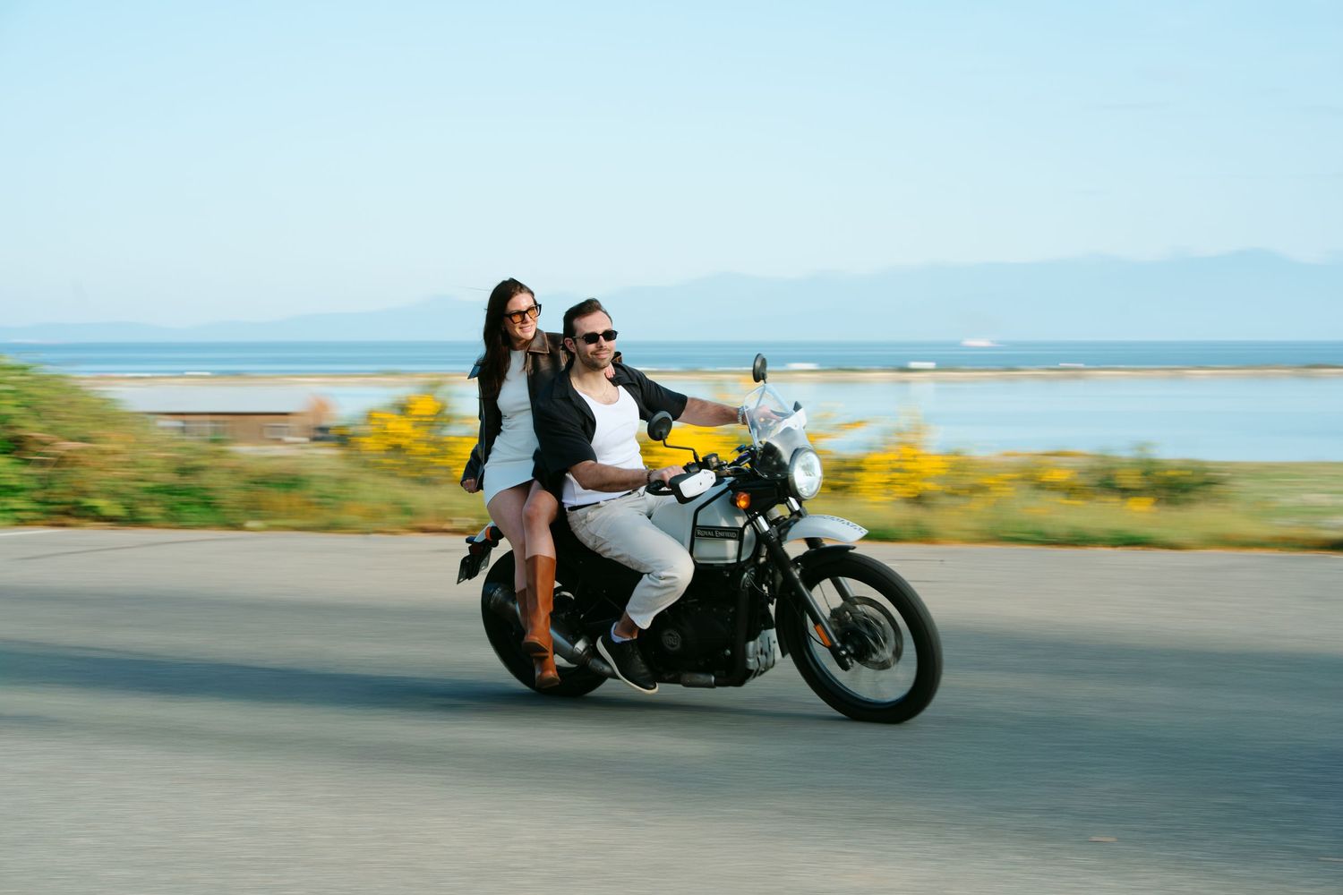 Couple riding a classic motorcycle on a scenic coastal road, woman in black jacket sitting behind man wearing sunglasses.