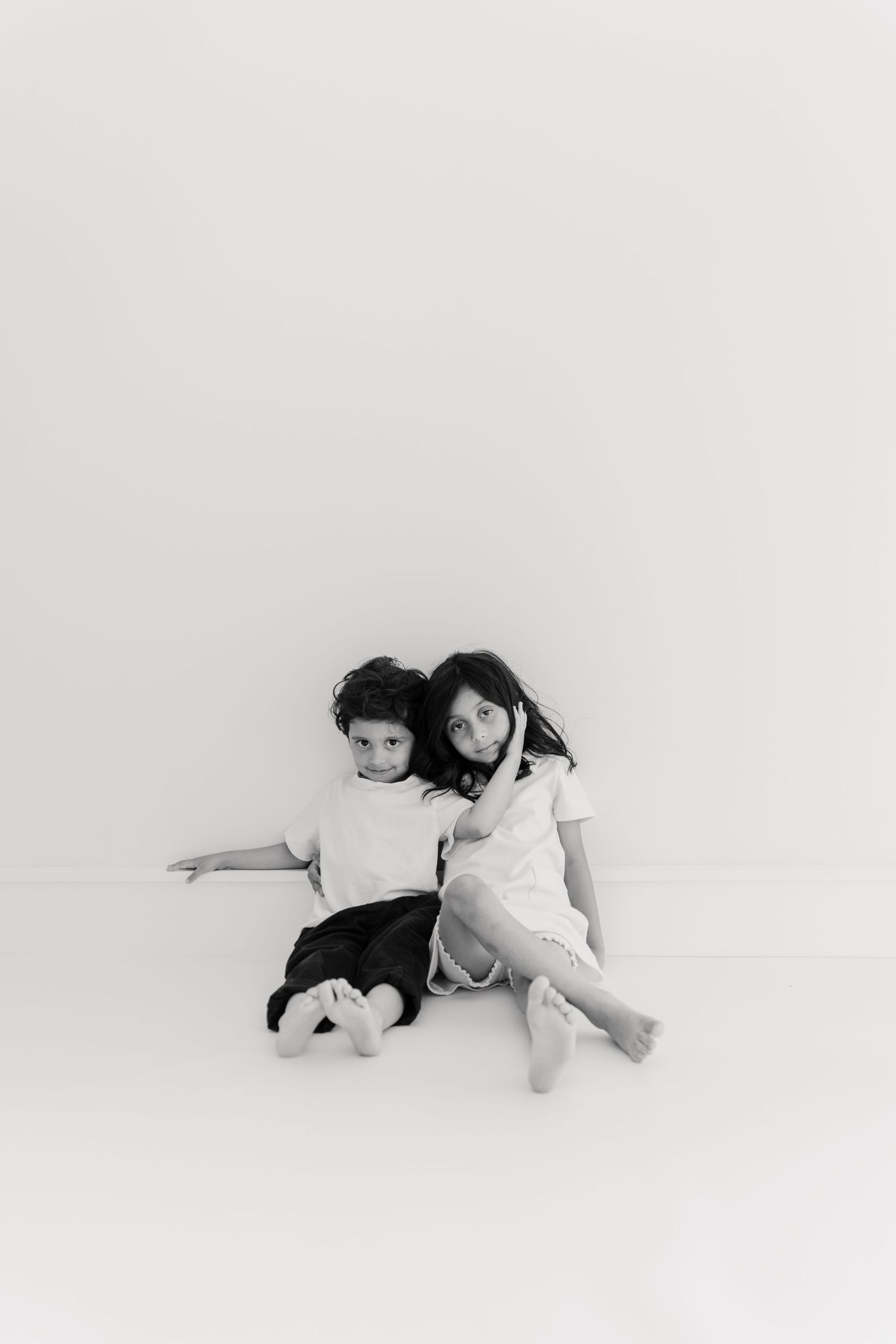 Two children sitting together against a white backdrop in a minimalist black and white portrait.