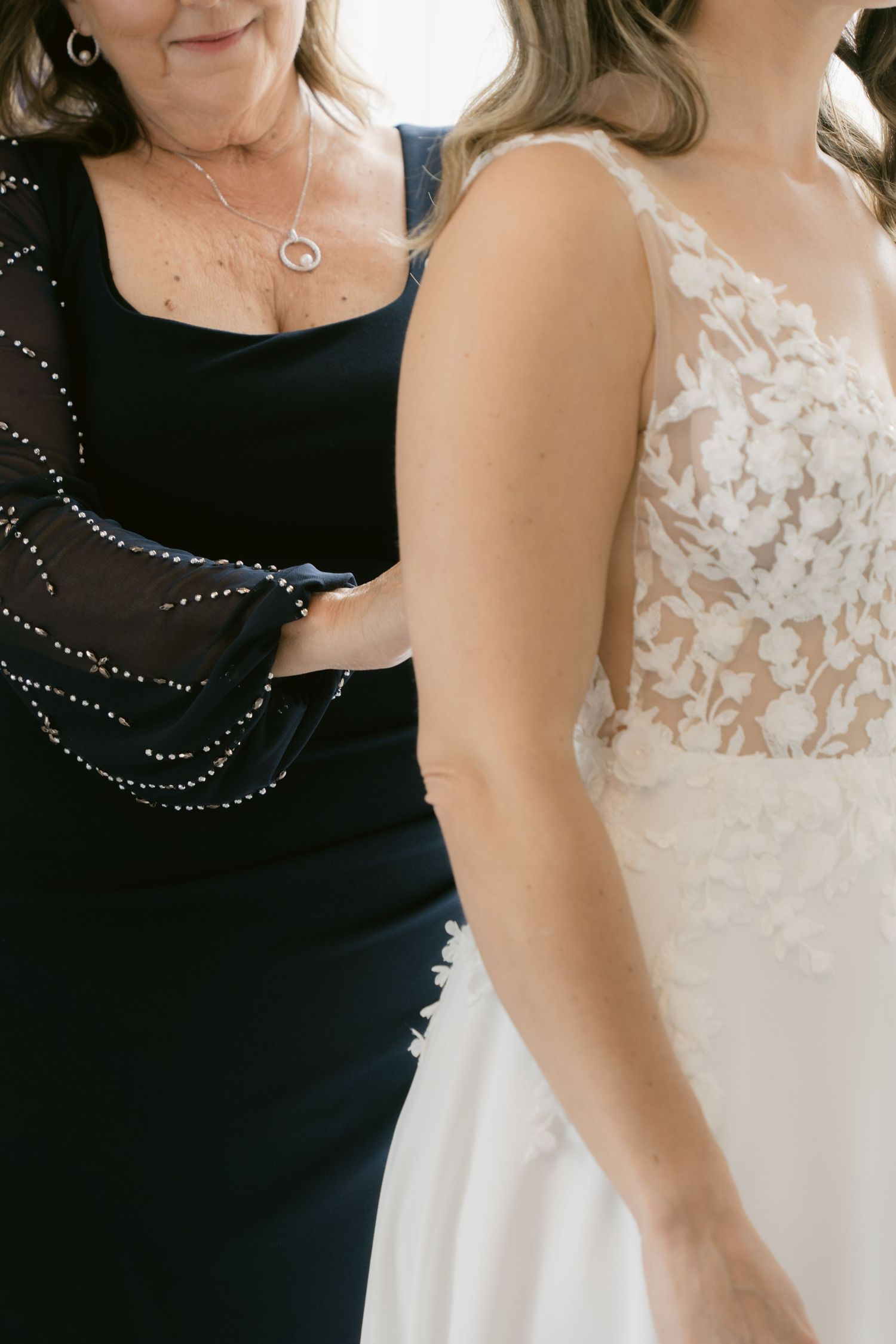 Detail shot of bride in lace wedding gown standing next to woman in navy dress with sparkle details.