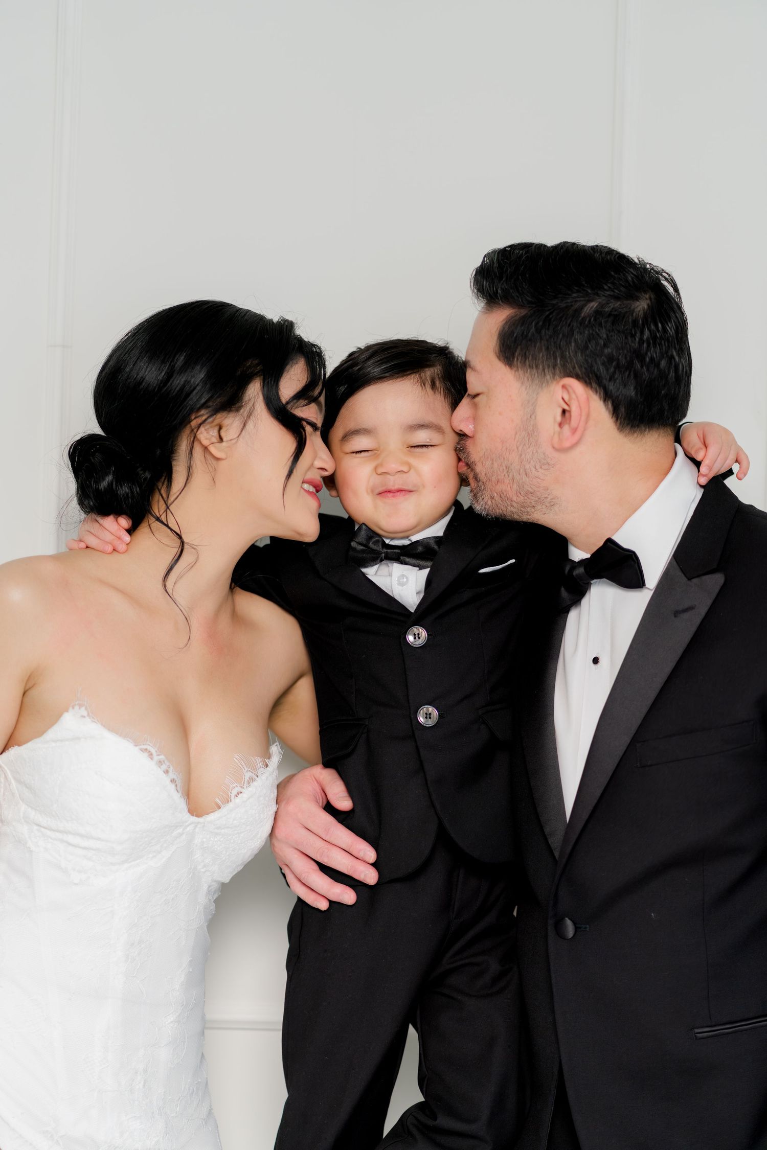 A bride and groom in formal wedding attire kiss their young son dressed in a matching black tuxedo.