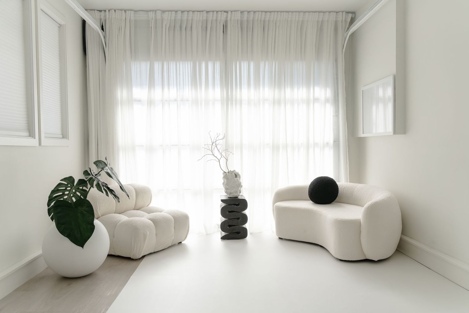 Modern white living room with curved sofas, plants and sheer curtains in bright natural light.