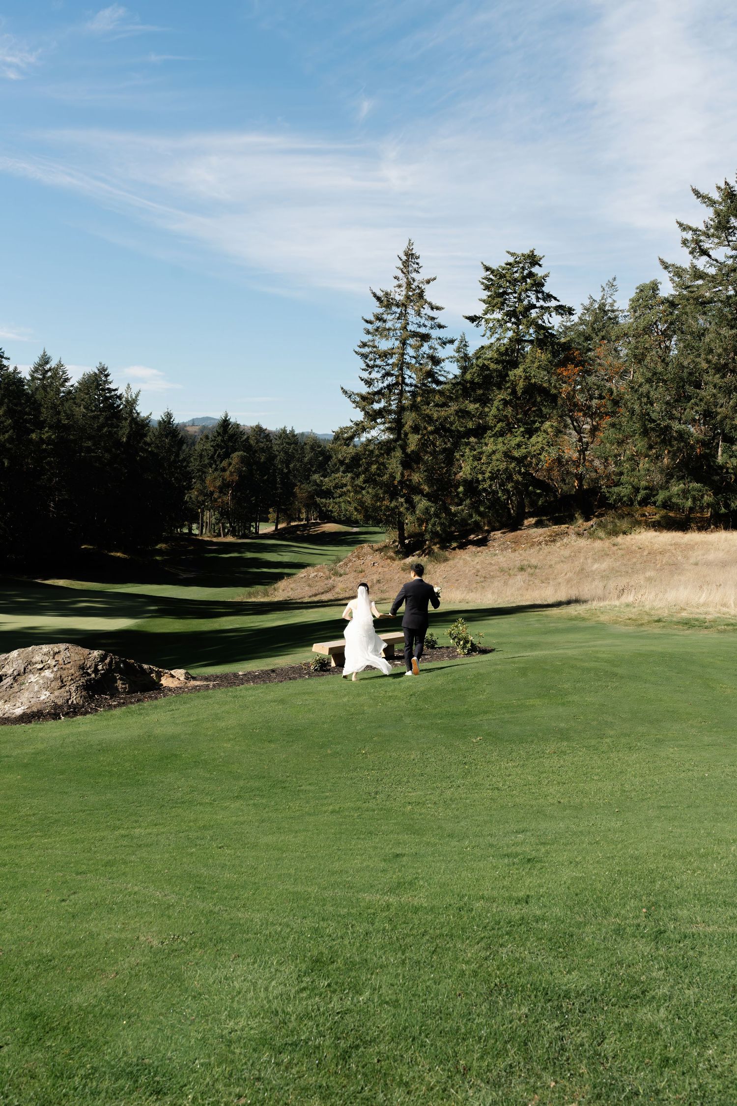 Bride and groom walking hand in hand across a lush green golf course surrounded by tall evergreen trees under a blue sky. Victoria BC