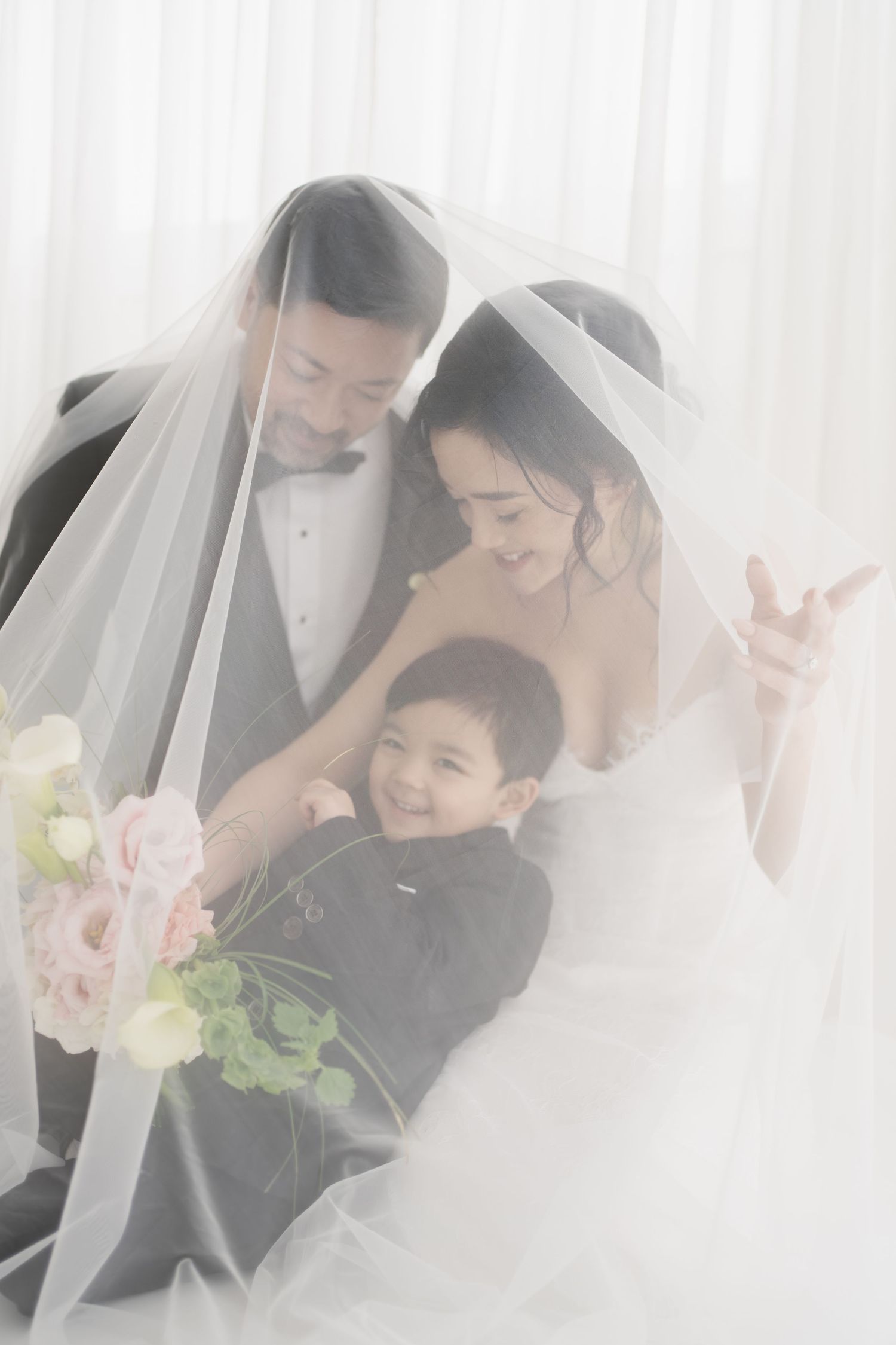 Bride and groom in wedding attire share a tender moment under a veil with a smiling young boy in a suit.