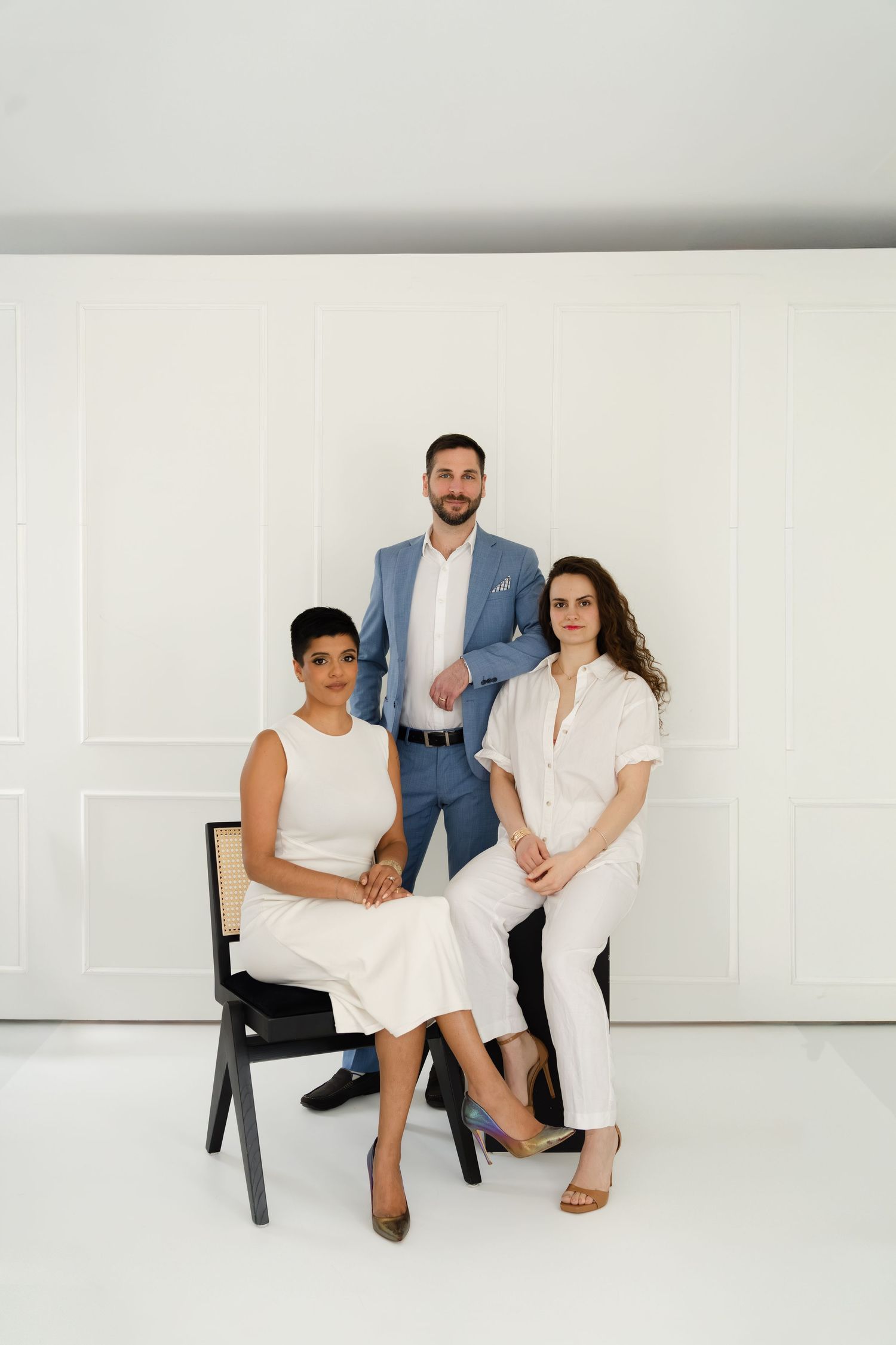 Three professionals pose in a bright white studio, two women seated and a man standing, wearing elegant neutral outfits.