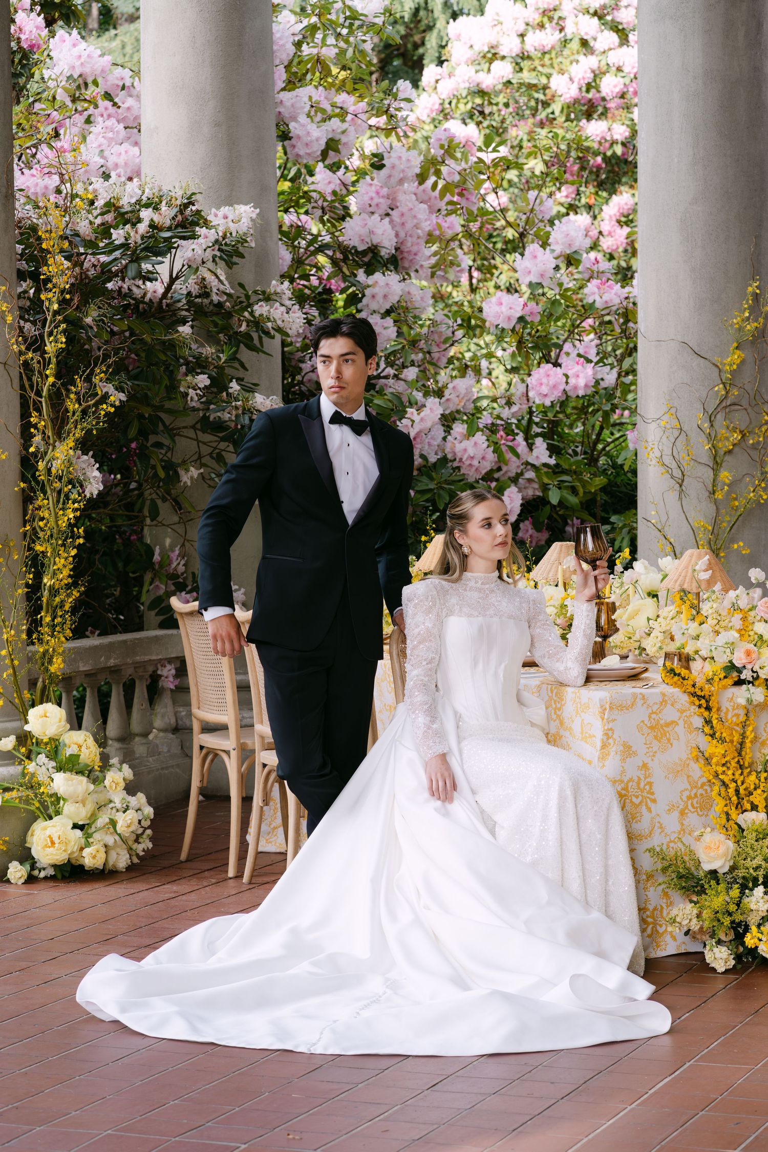 Bride in white lace gown seated beside groom in black tuxedo, surrounded by blooming pink flowers and yellow roses.