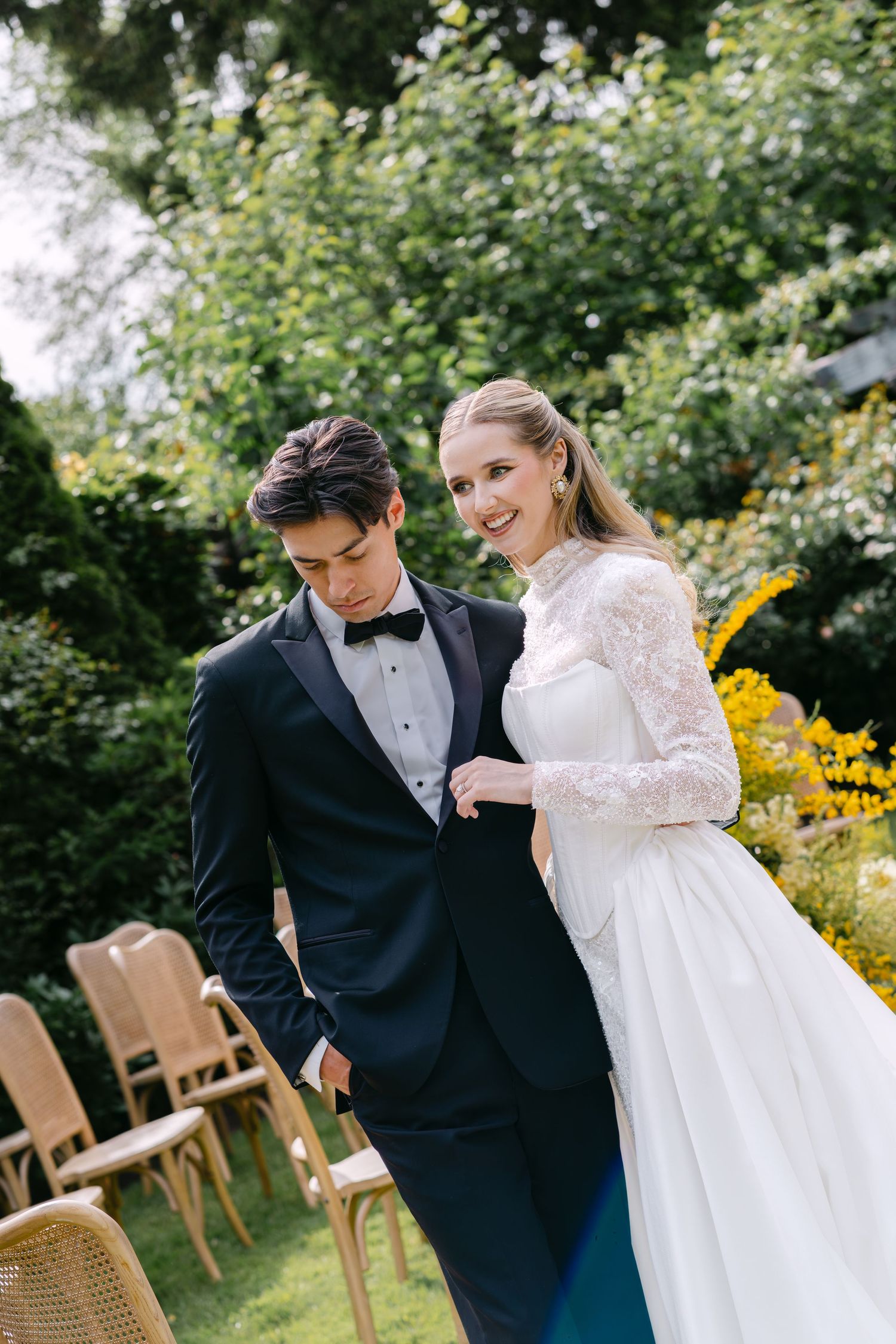 Bride in lace wedding gown smiles beside groom in black tuxedo at outdoor garden wedding ceremony.
