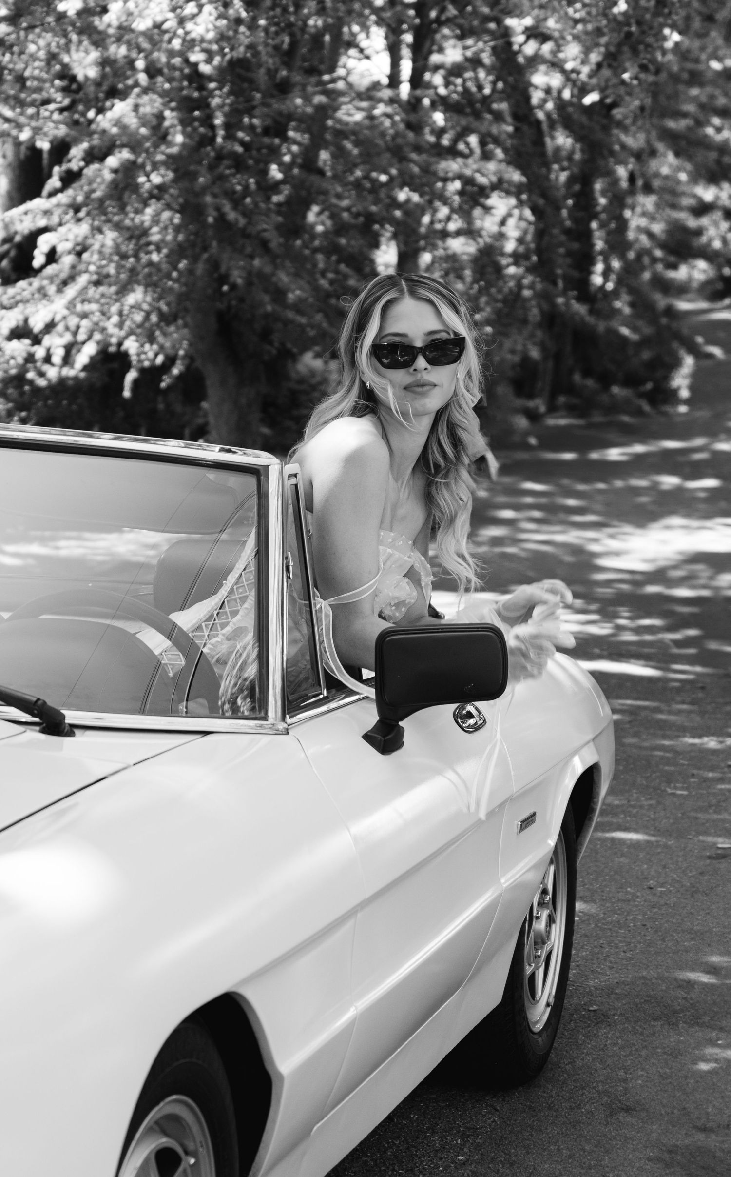 Classic white convertible car photographed in black and white with sunlit trees.
