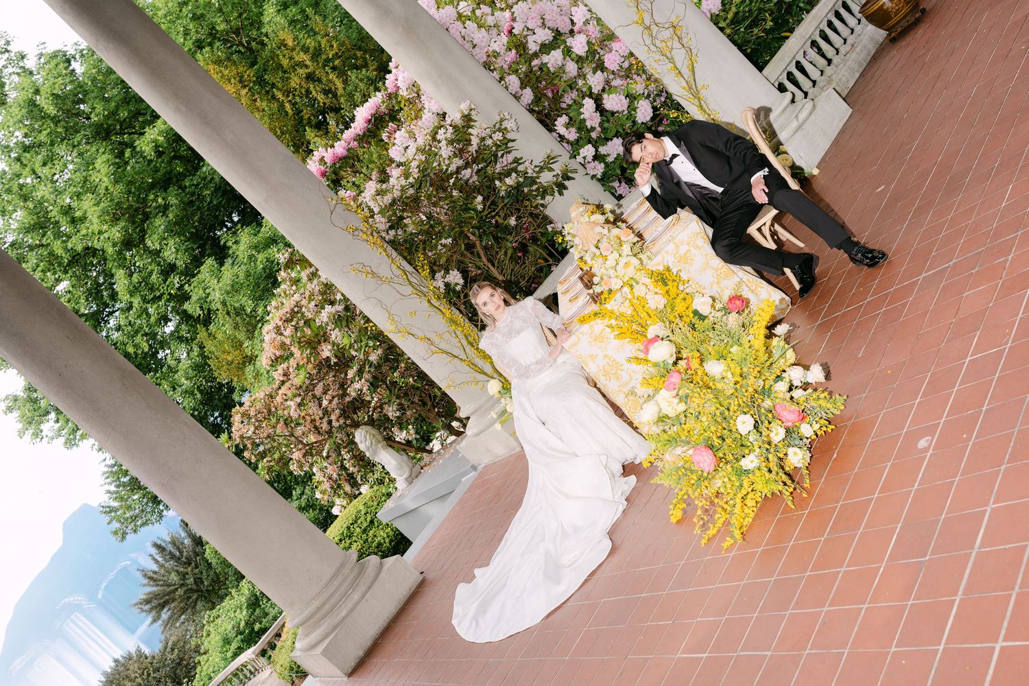Aerial view of wedding couple kissing amid colorful floral arrangements on a sunlit outdoor terrace.