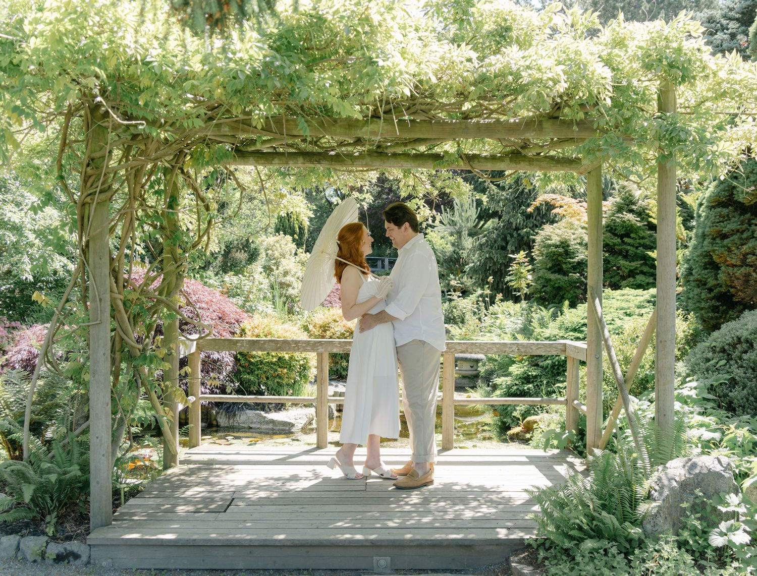 A couple in white outfits share an intimate moment on a covered porch with sunlit garden backdrop at Japanese Garden located in Gorge park