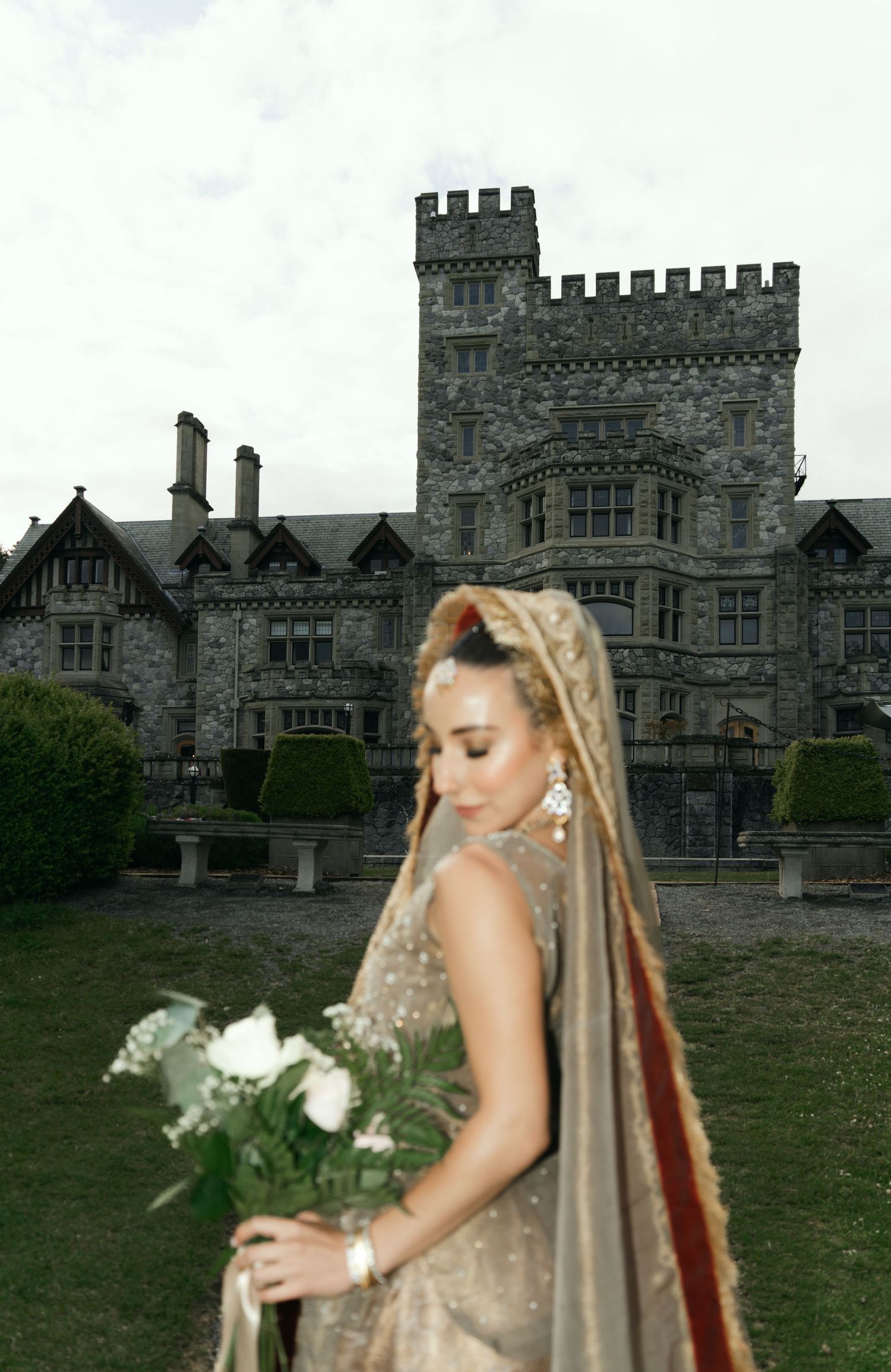Bride in gold embroidered lehenga holding white bouquet stands before a grand stone castle. Victoria BC