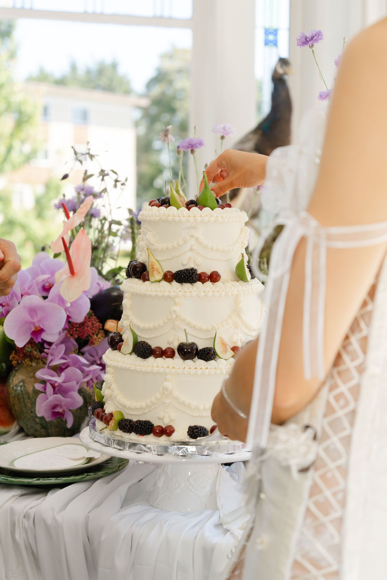 Elegant three-tier white wedding cake adorned with fresh berries and flowers on a crystal stand beside a bride in white dress.