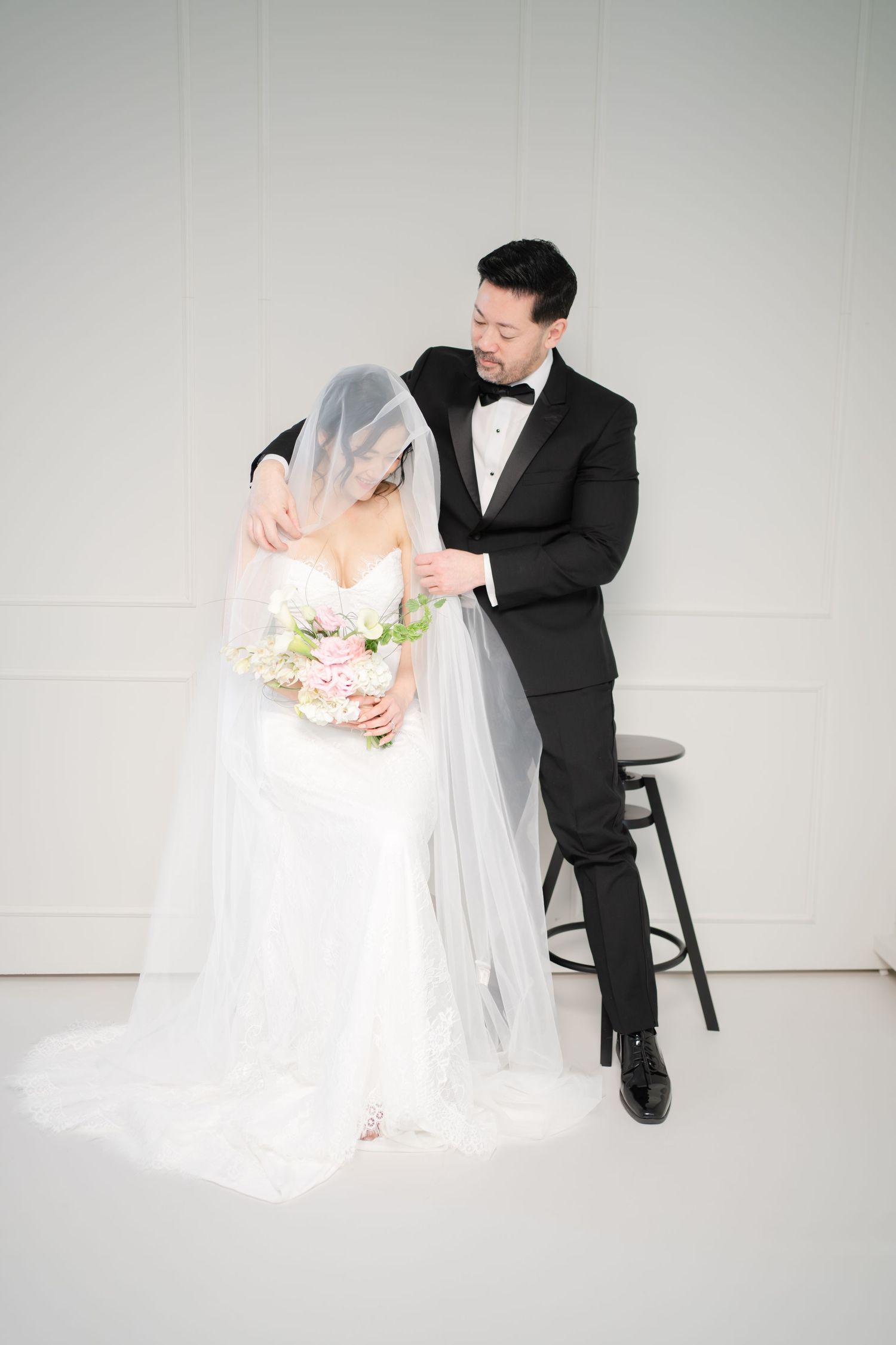Groom in black tuxedo lifting bride's veil while she holds pink floral bouquet in white wedding dress.