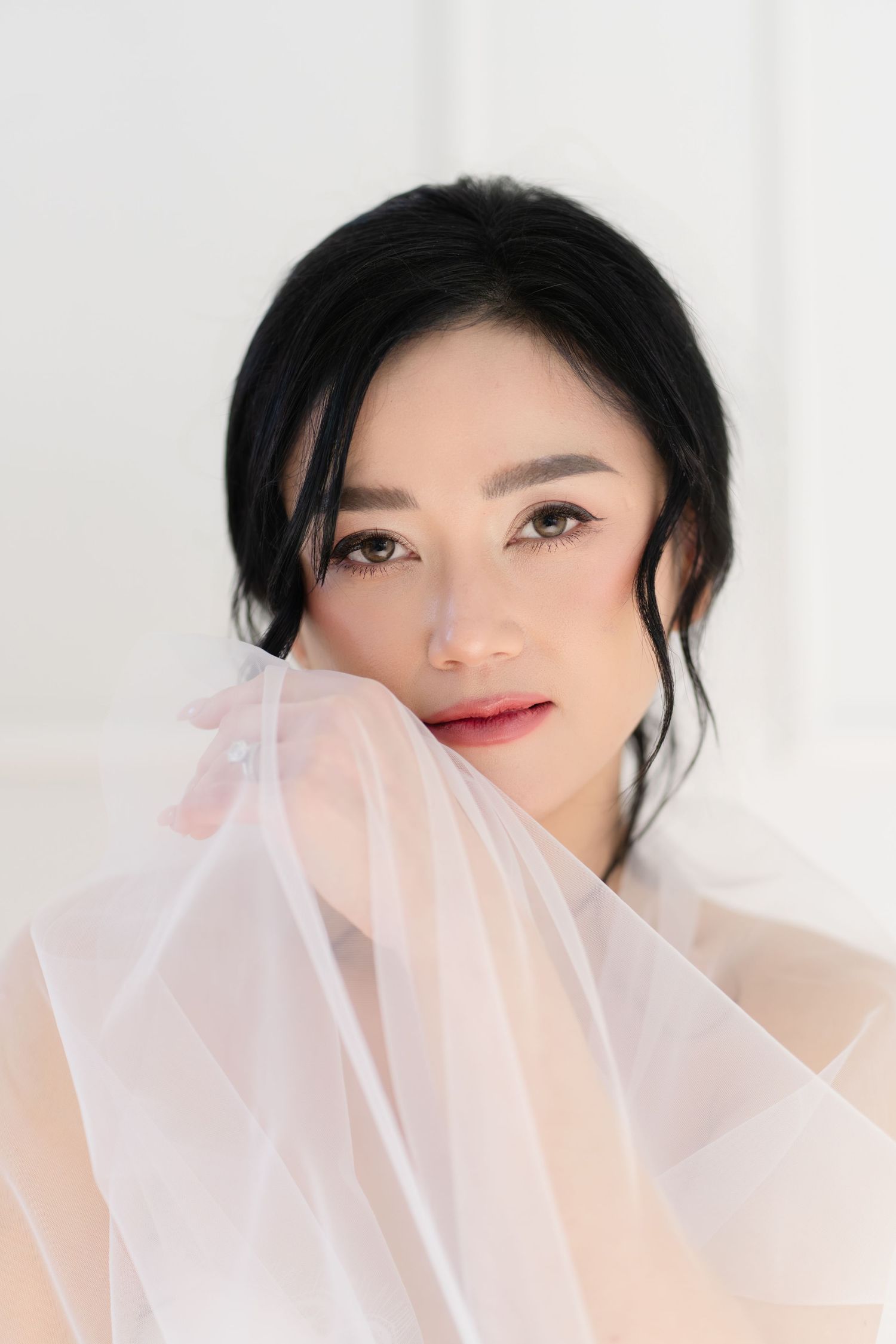 A beautiful Asian woman with dark upswept hair and elegant makeup poses gracefully behind a sheer pink veil.