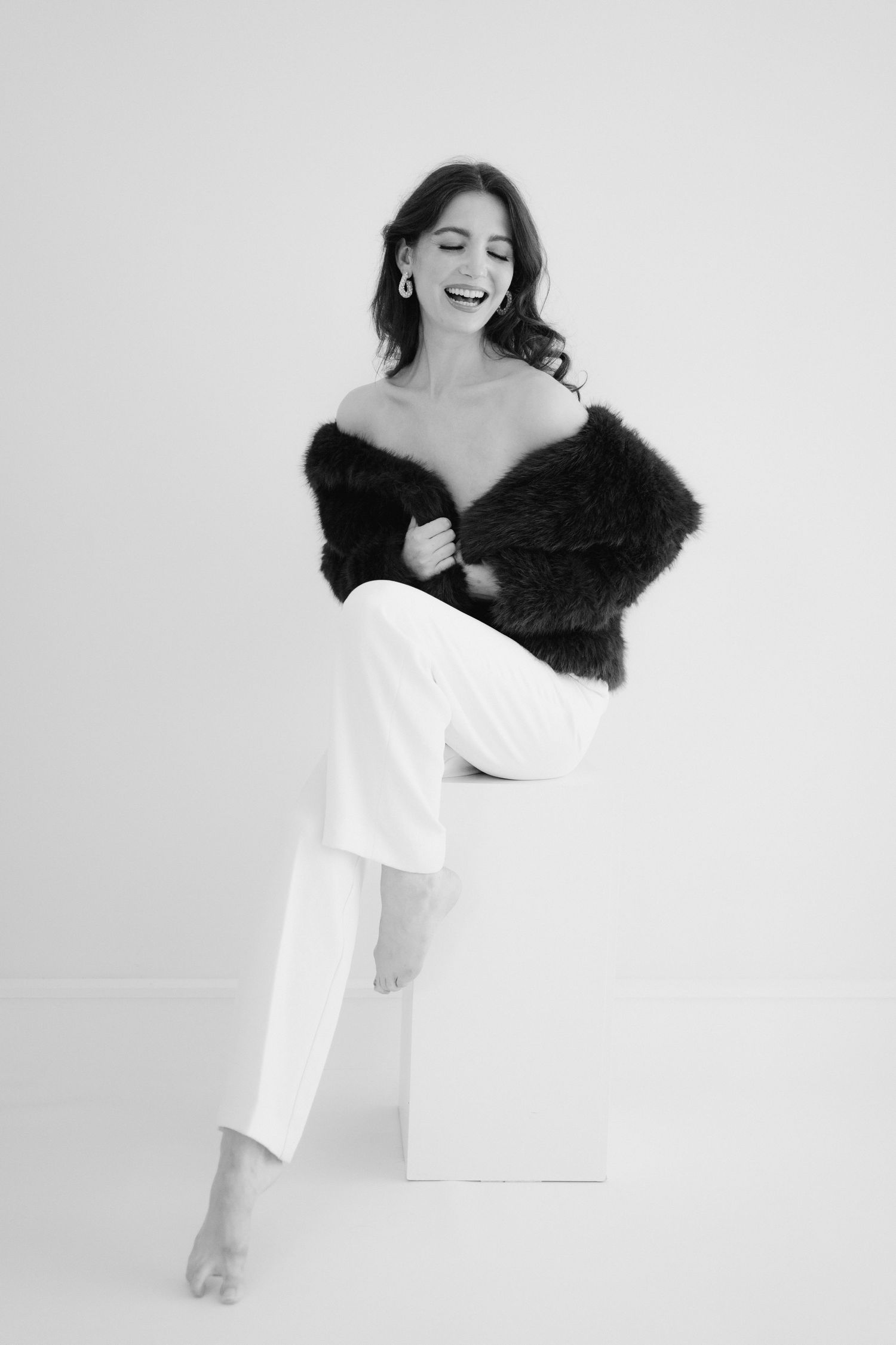 A figure in a white outfit poses elegantly with a black fur stole in a minimalist studio setting.