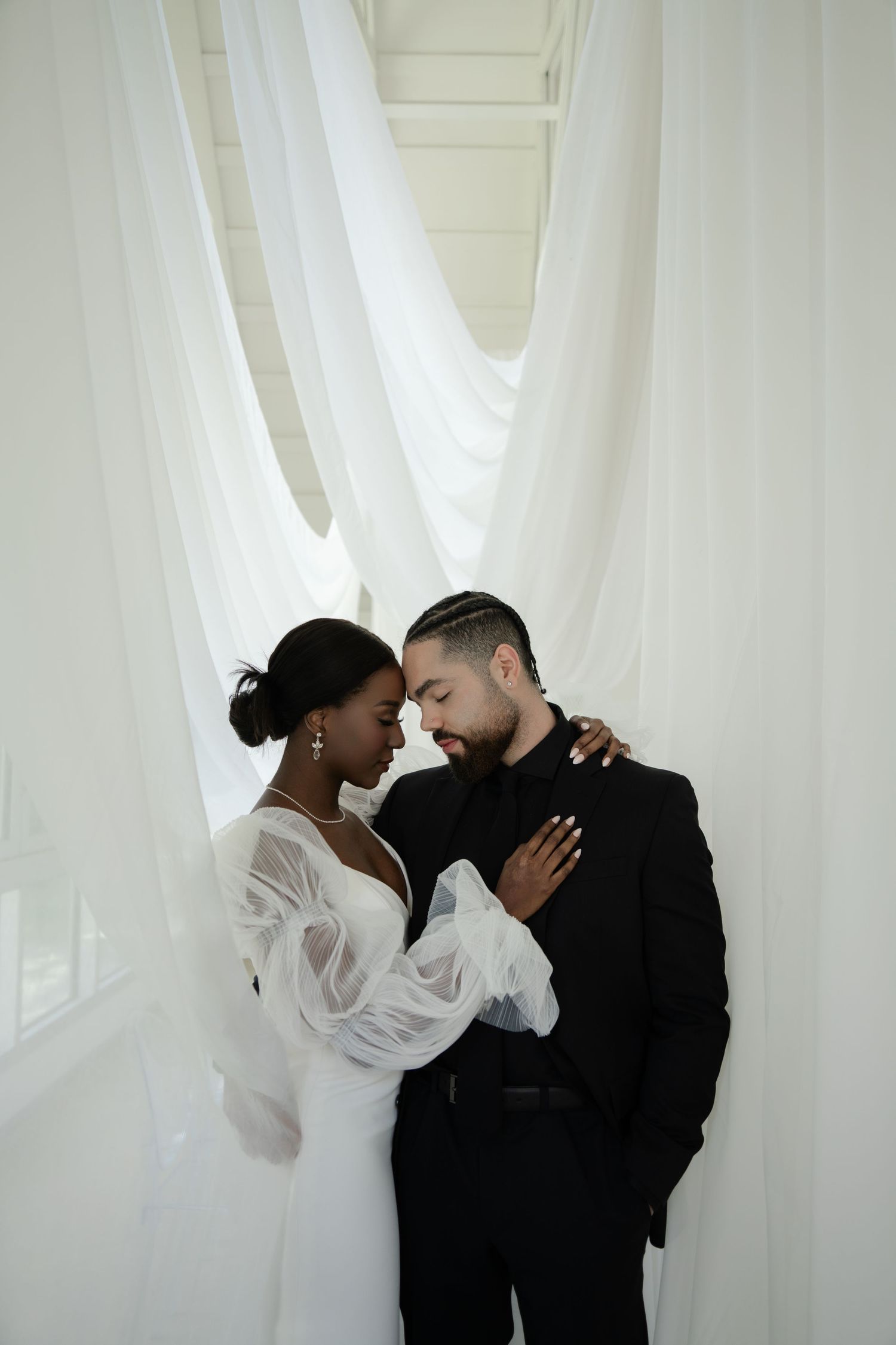 A Black bride in a white gown and groom in black suit share an intimate moment against flowing white drapes.