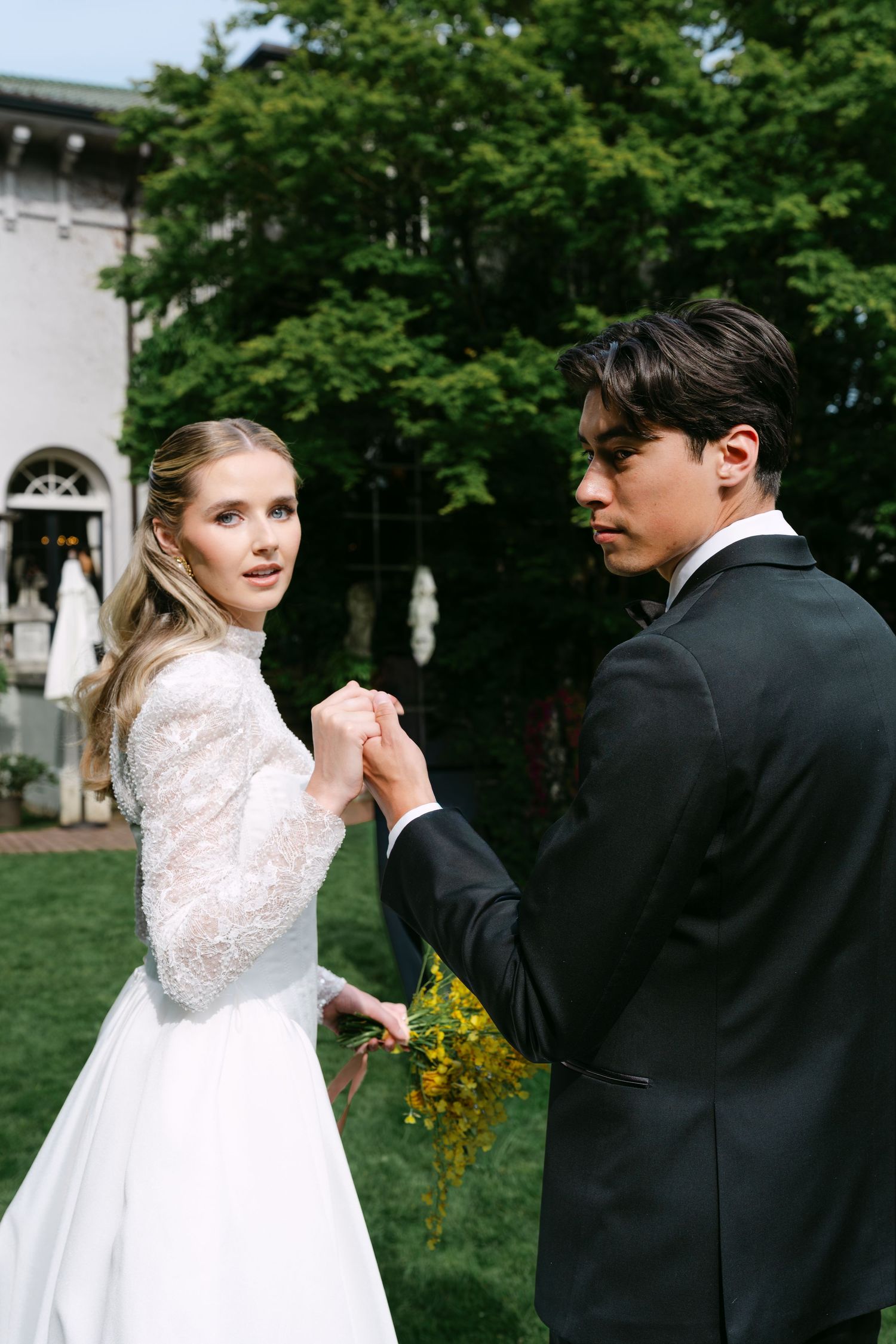 Bride and groom hold hands outdoors, she in lace wedding gown, he in black tuxedo, dancing in a Hycroft Manor garden.