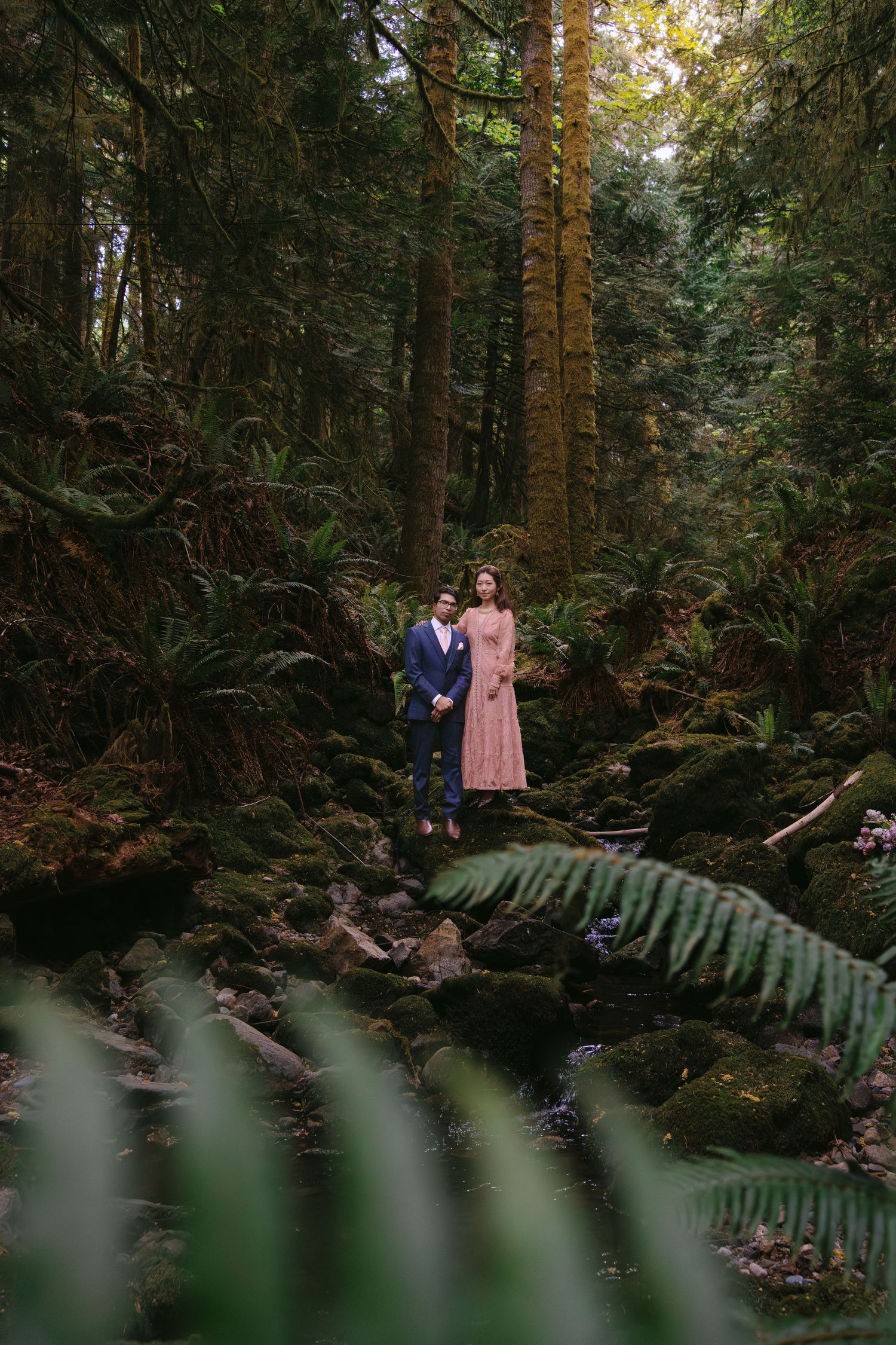 Couple in formal attire standing on mossy rocks in a lush, fern-filled forest setting at Cedar Haven Sooke BC