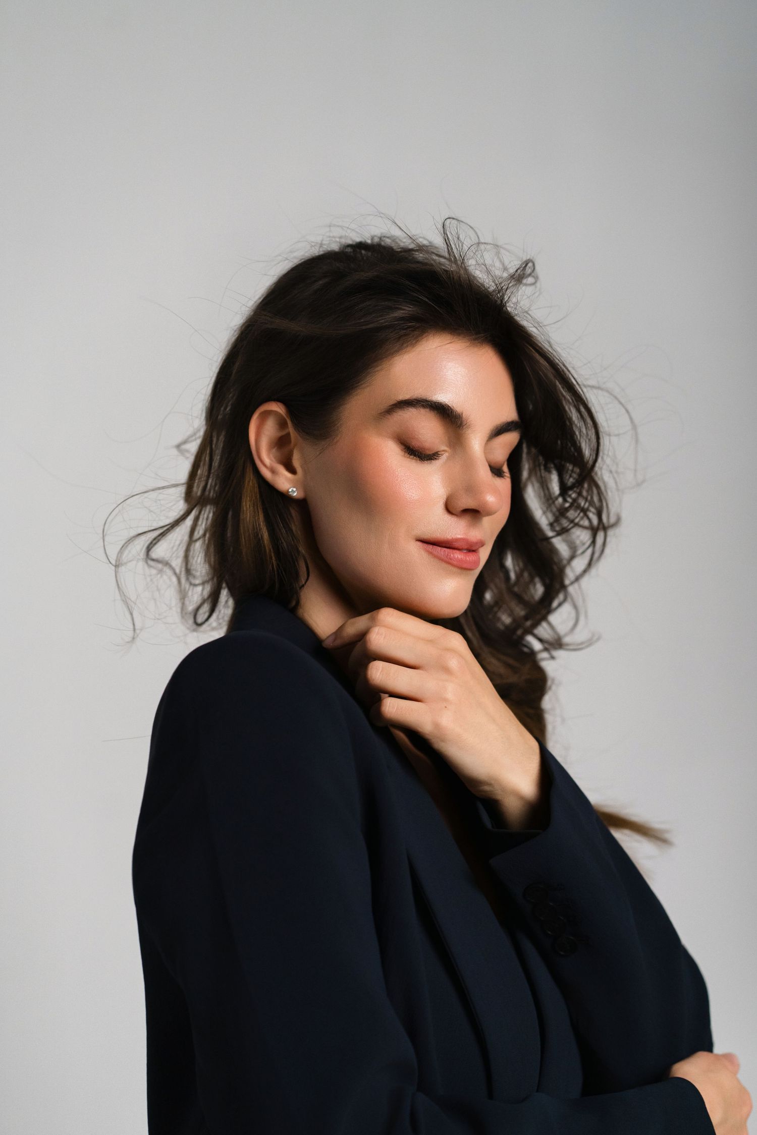 A young woman in a navy blazer smiles serenely with closed eyes, her dark wavy hair flowing against a gray background.