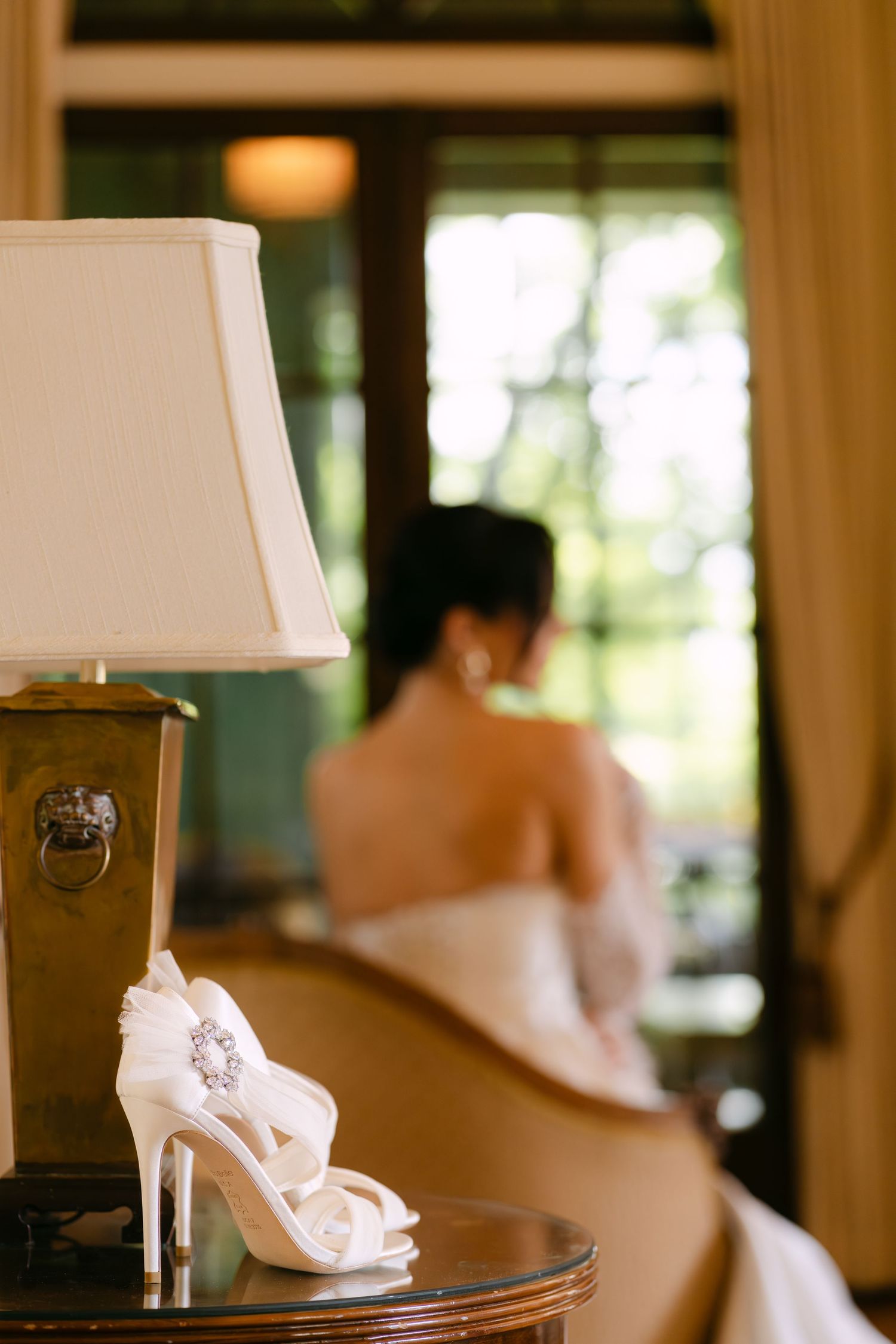 White bridal heels with crystal brooch on table beside lamp, bride in wedding dress seated in background.