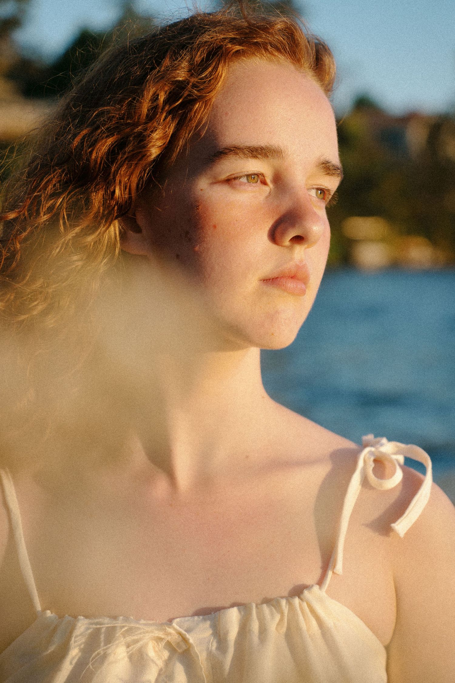 Young redhead woman with freckles in white sundress gazing into distance by sunlit waterfront at golden hour.