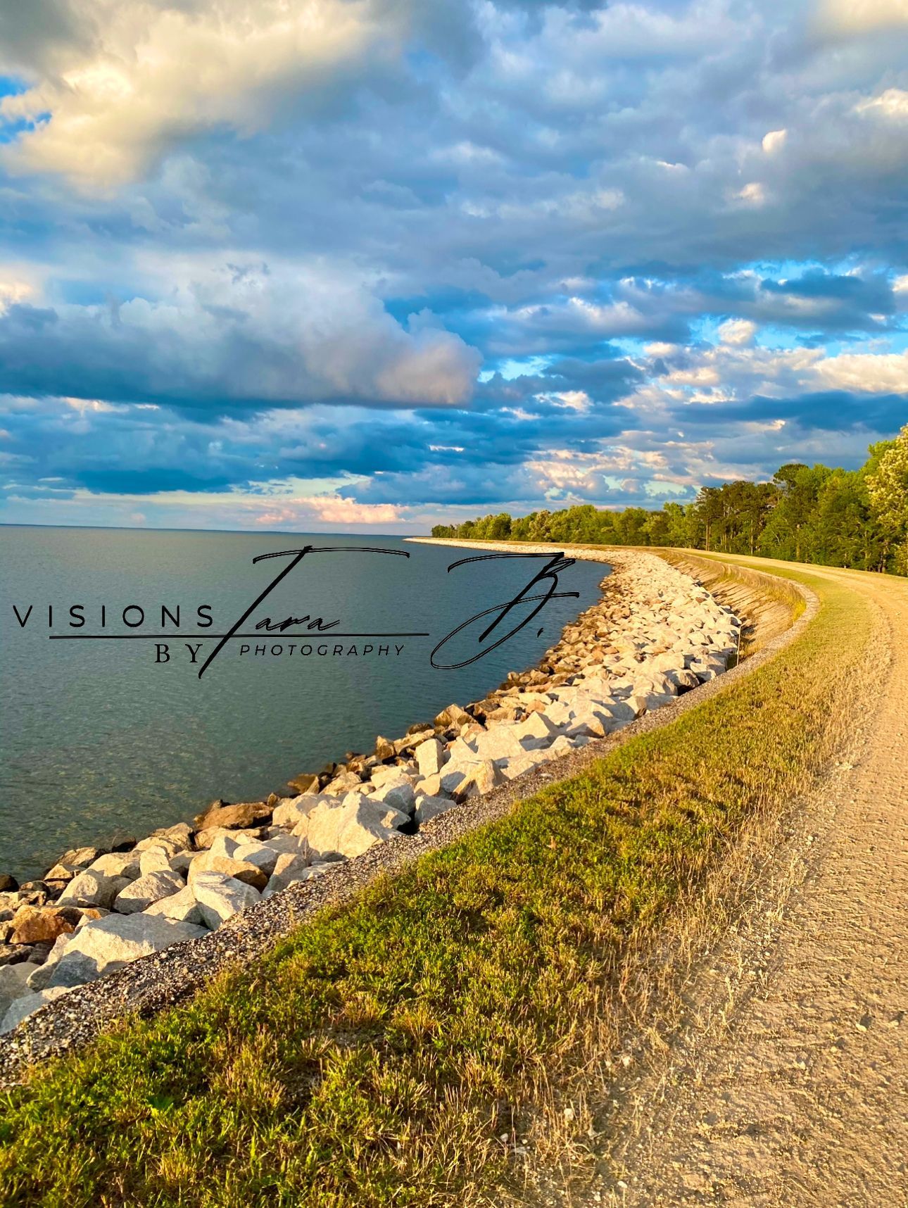 Curved lakeside path leading toward water and mountains under a blue sky with clouds, capturing a peaceful scenic outdoor view.