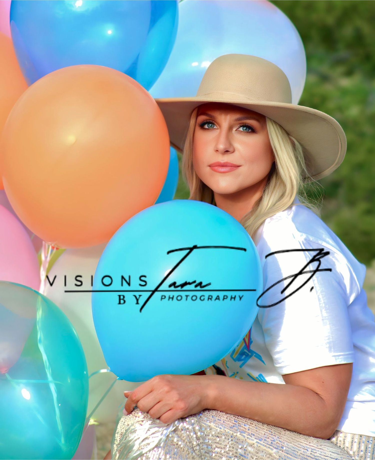 Woman holding colorful balloons while wearing a hat, looking softly toward the camera with a radiant expression.