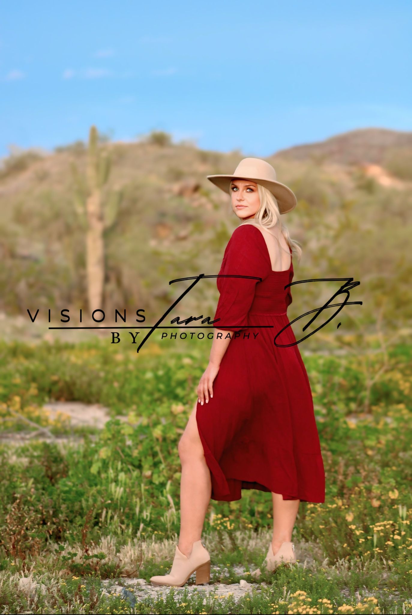 Woman in a red dress walking through desert wildflowers, glancing back with mountains and blue sky behind her.