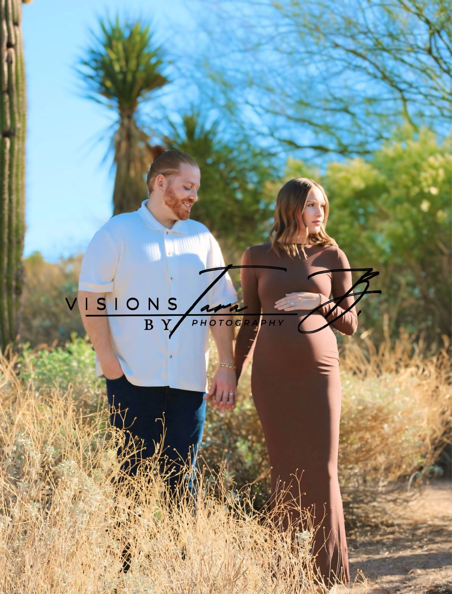 Pregnant couple standing together in the desert, holding hands and smiling in warm sunlight surrounded by natural scenery.