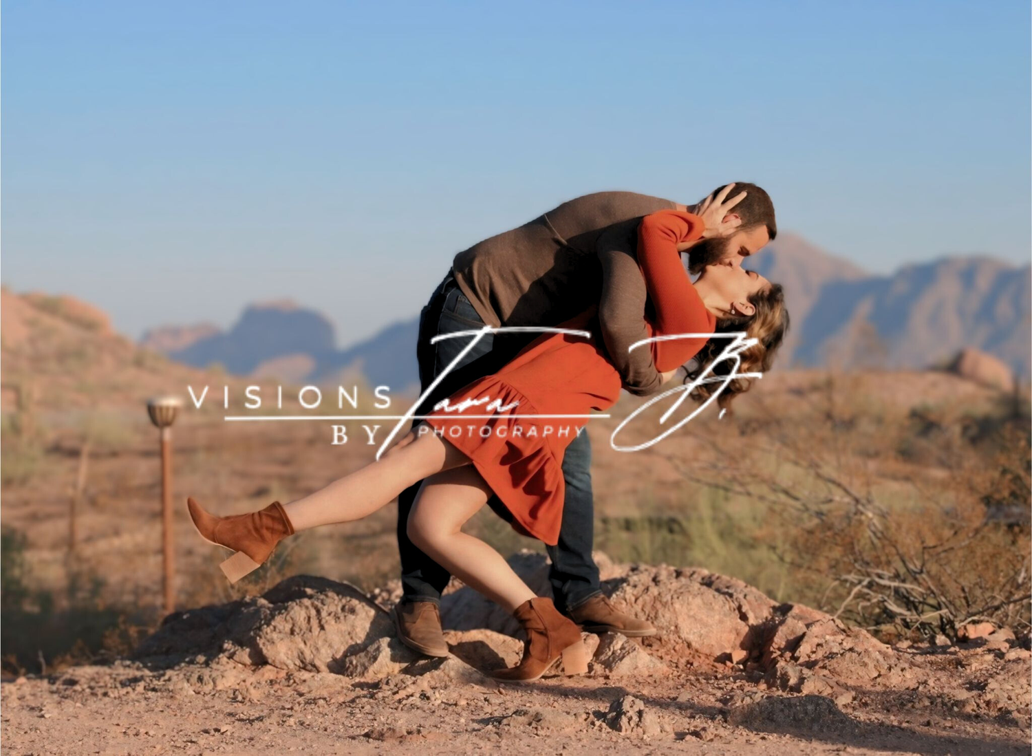 Couple sharing a romantic dip and kiss in the desert, surrounded by warm sunlight and mountain views.