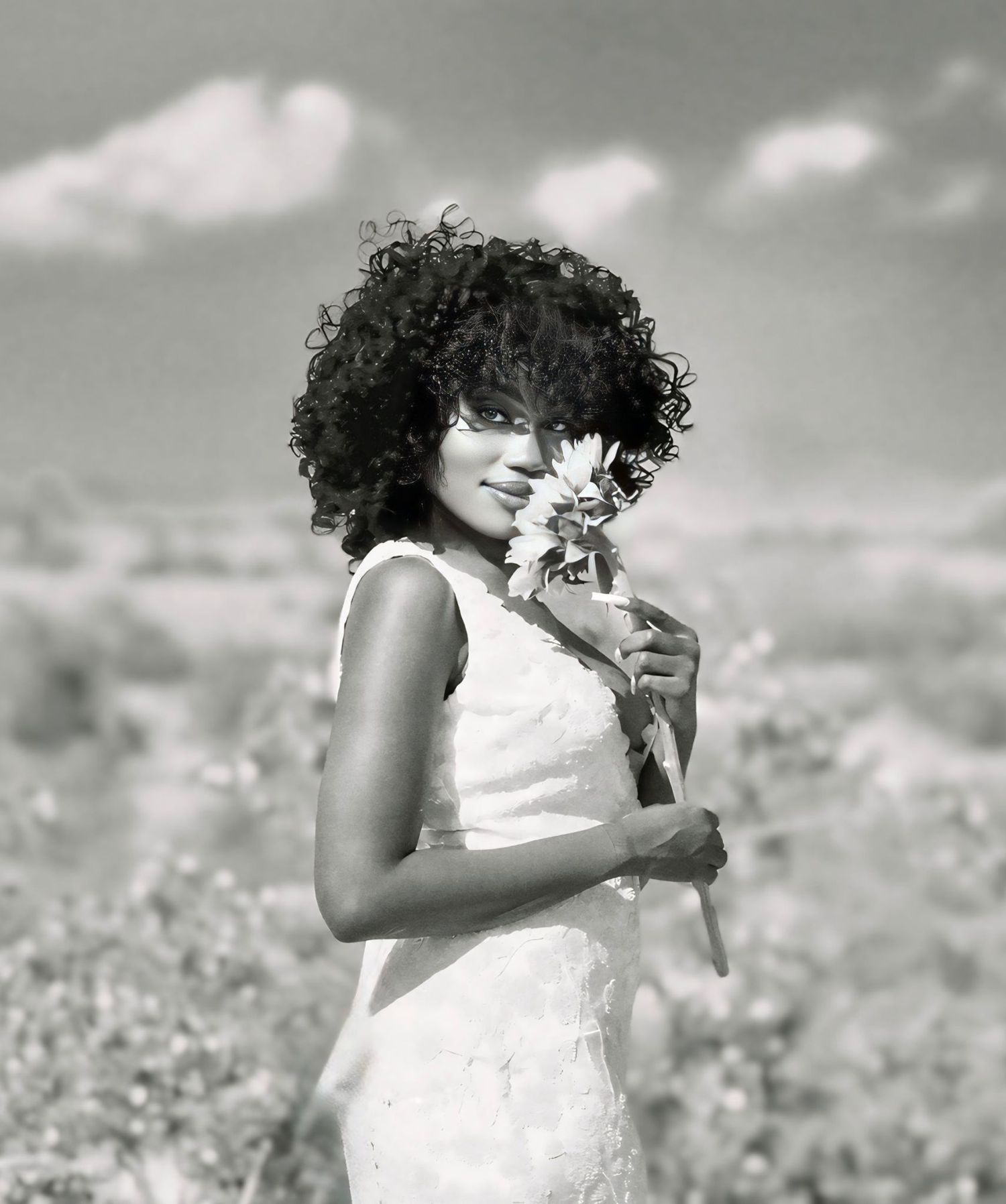 Black-and-white portrait of a woman smiling gently outdoors, holding her dress as she stands in soft natural light.