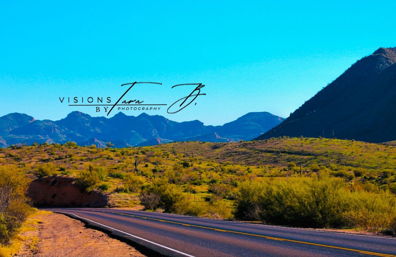 Open desert road leading toward bright green hills and blue mountains under a clear sky, capturing a peaceful scenic drive.