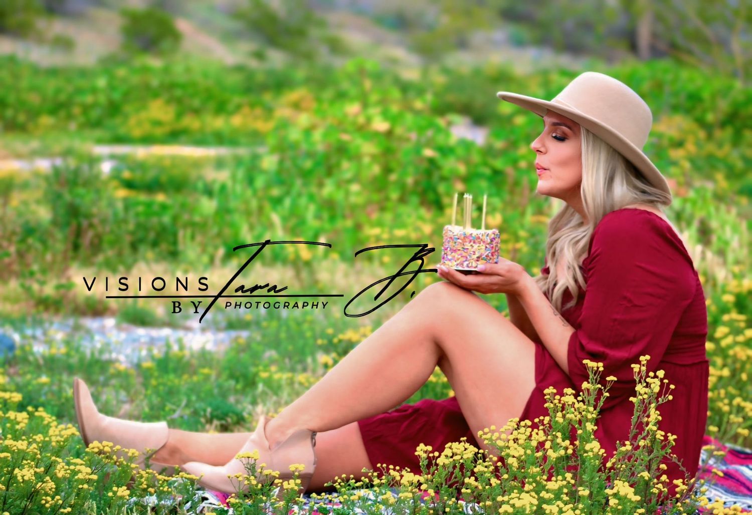 Woman sitting in blooming wildflowers wearing a hat and red dress, smiling while celebrating a peaceful birthday moment outdoors.