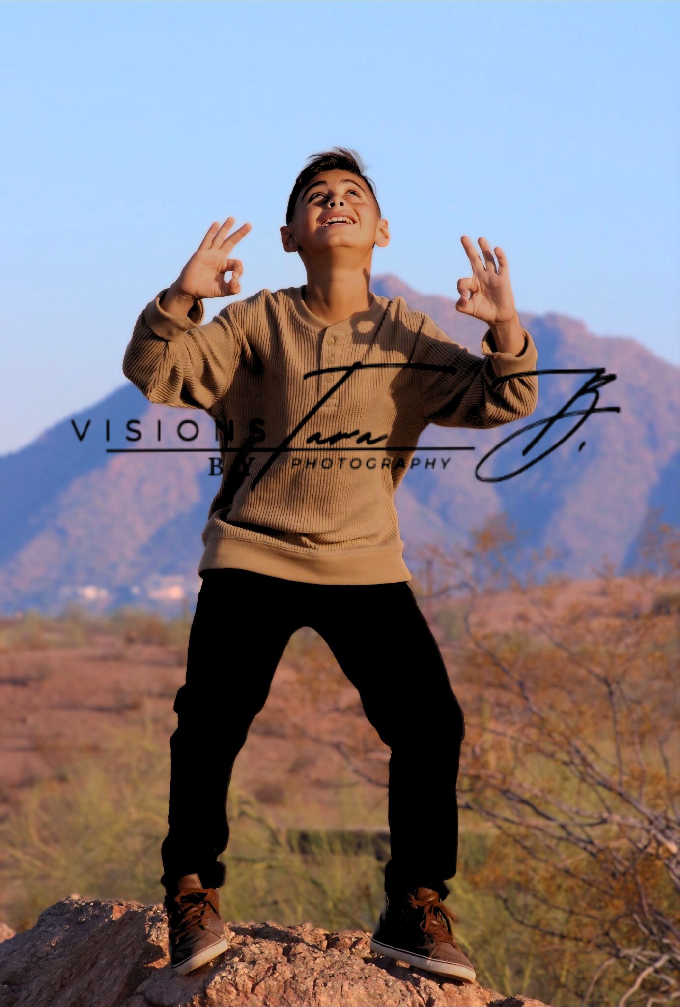 Boy posing confidently in the desert with sunlight behind him, standing in front of mountains and warm natural scenery.