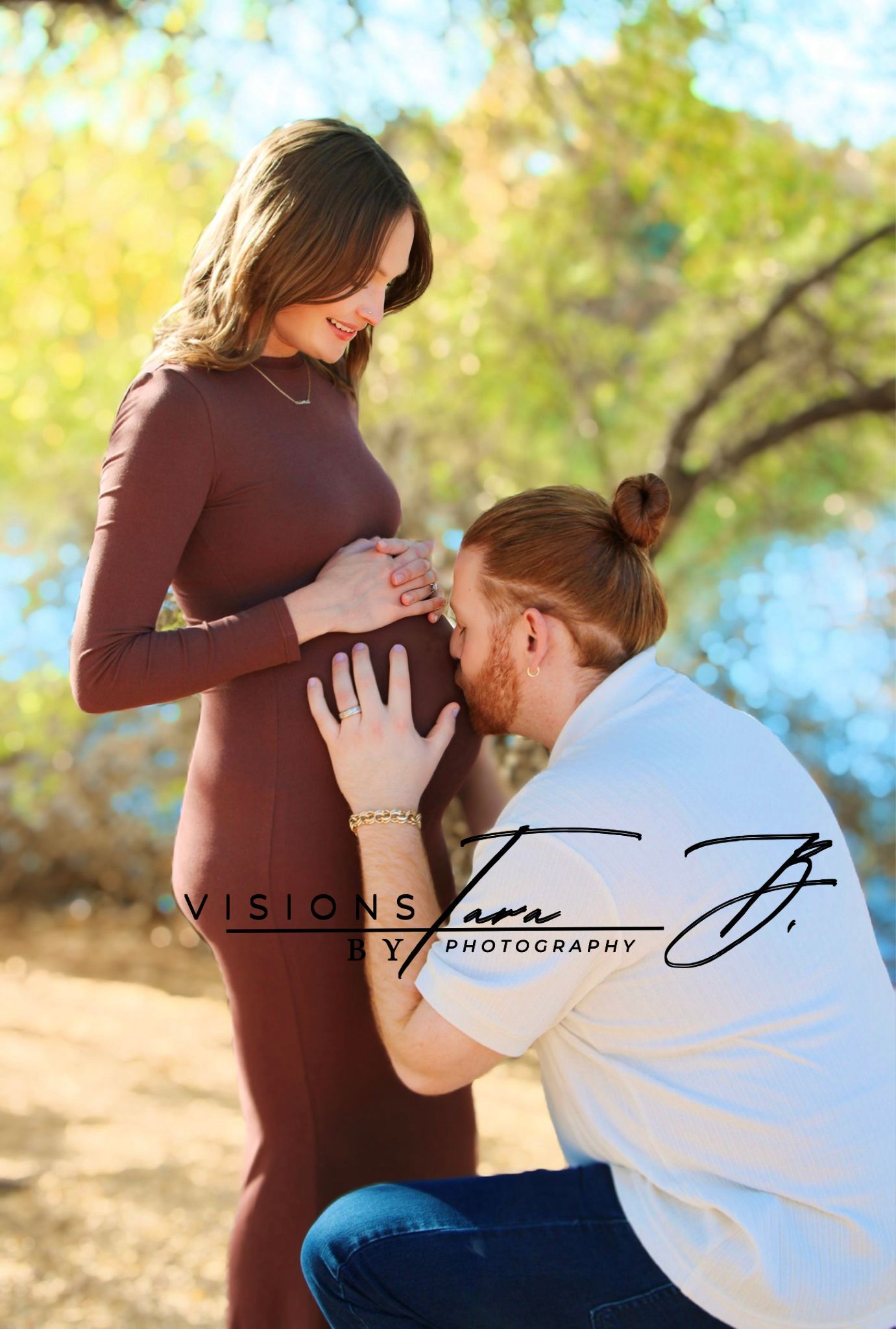 Partner kneeling to kiss the baby bump while the mother smiles softly, sharing a tender maternity moment by the water.