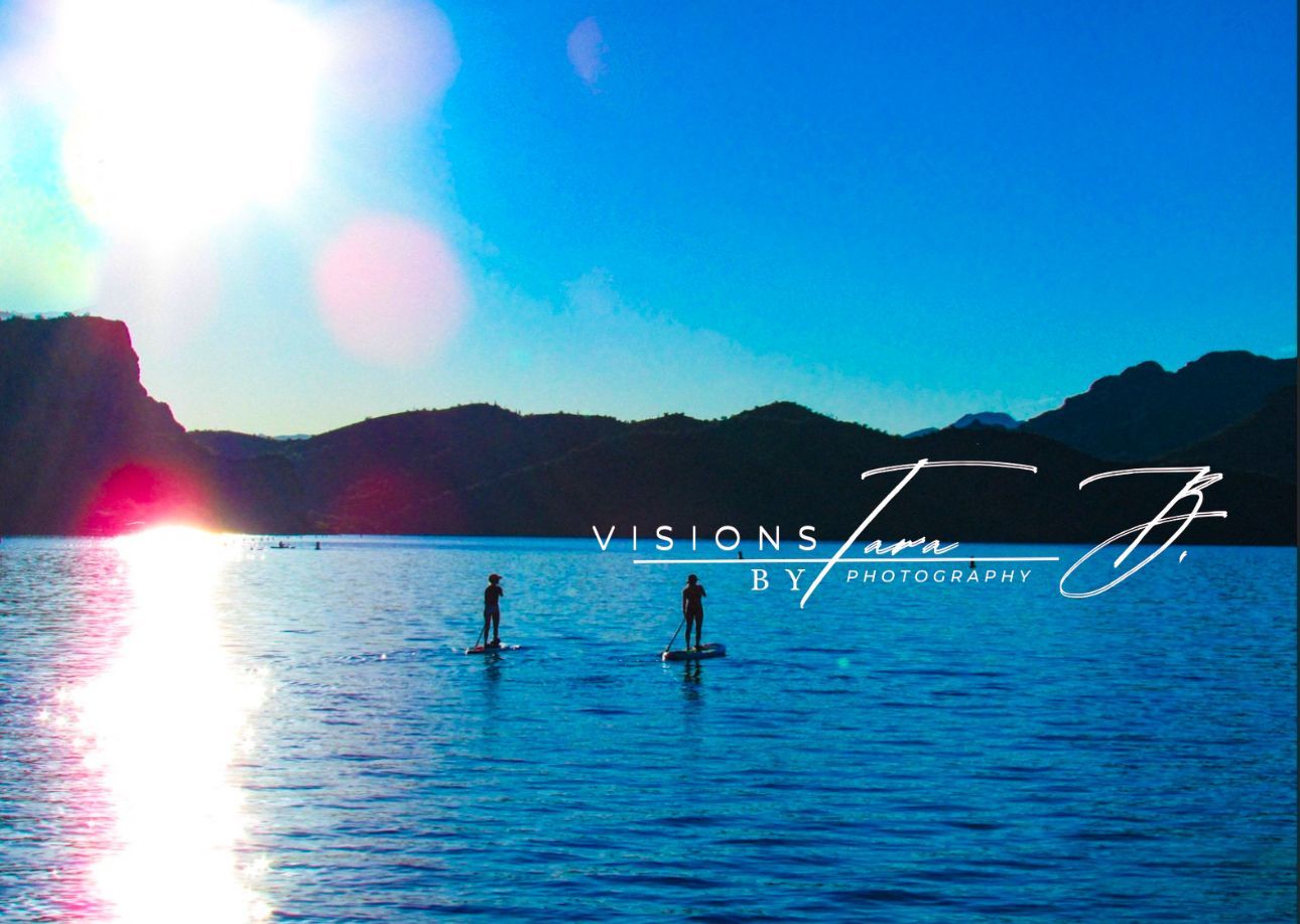 Two people paddleboarding on a bright blue lake with sun flares and mountains in the distance under a clear sky.