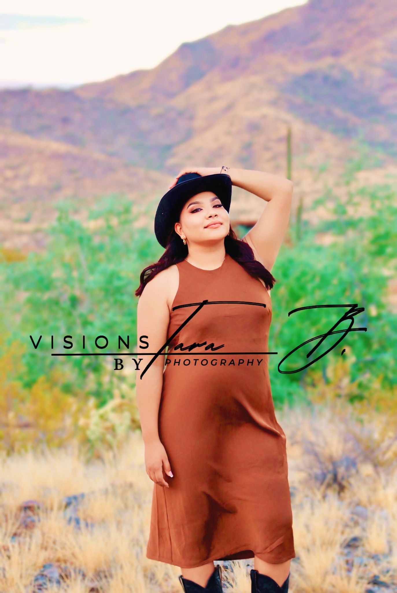 Woman in a cowgirl hat posing confidently in the desert, standing against sunlit mountains and natural scenery.