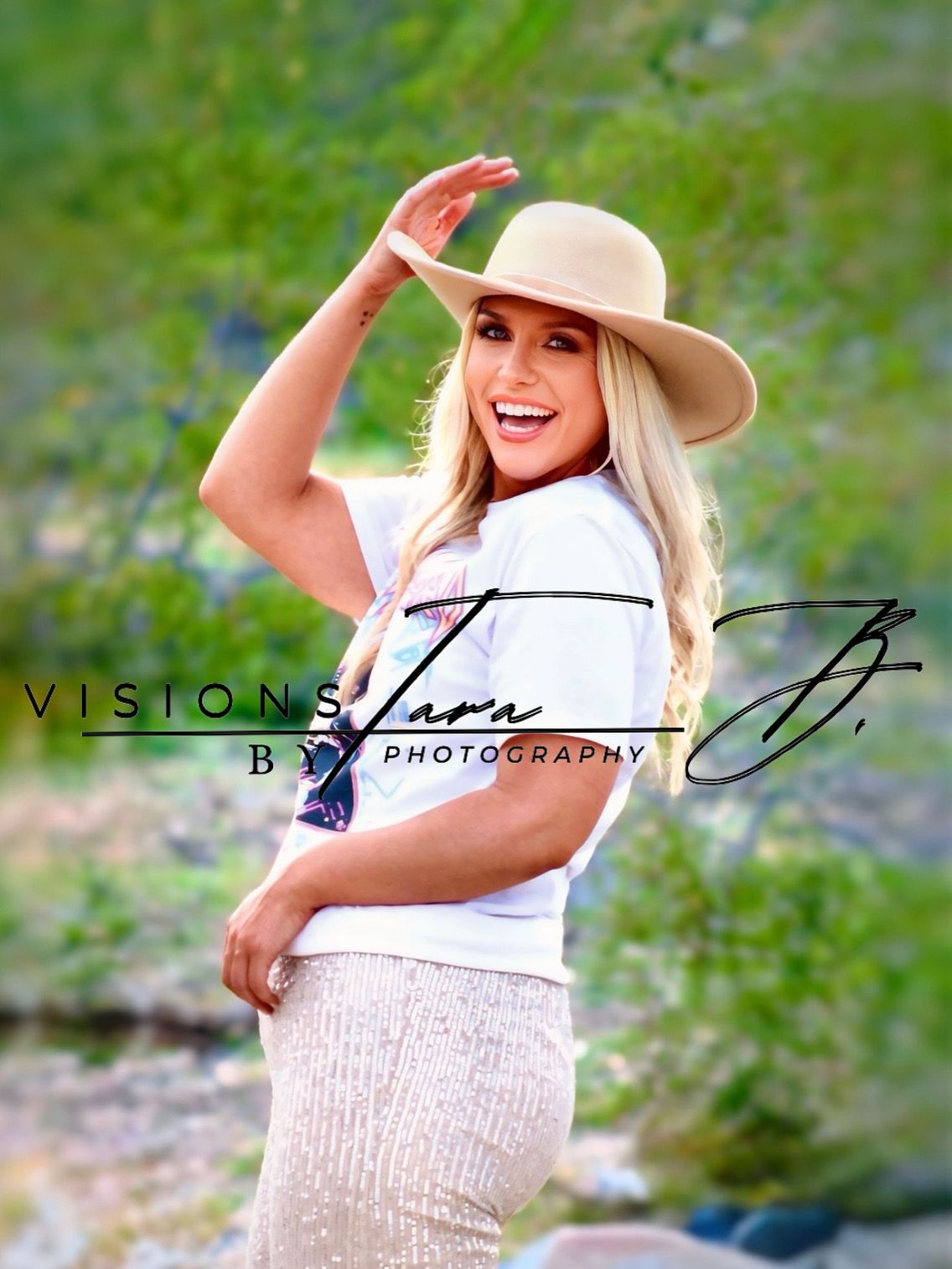 Smiling woman in a wide-brim hat posing in natural greenery, holding the brim as she stands in soft outdoor light.