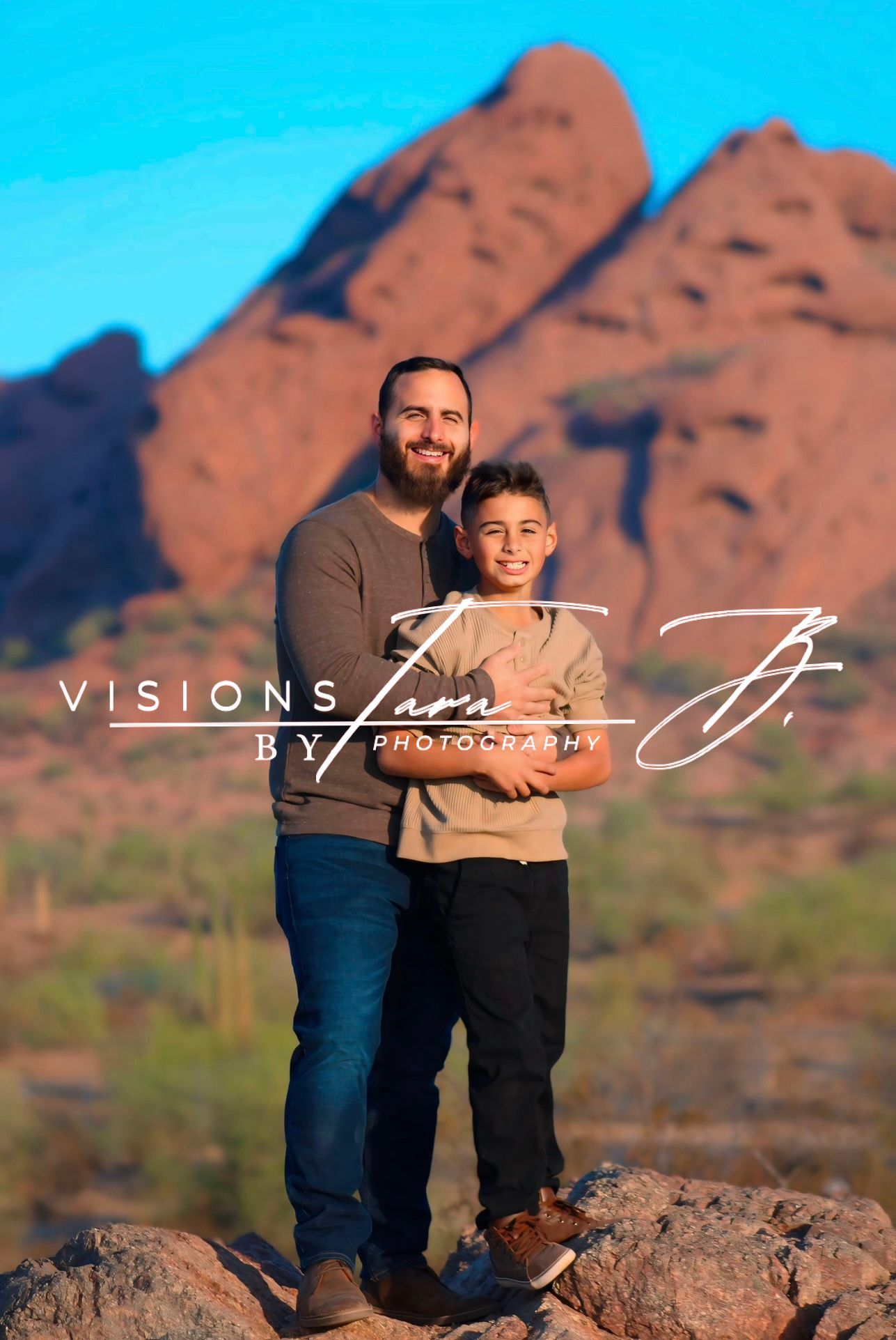 Father and son standing together in the desert with red mountains behind them, smiling during a warm outdoor portrait.