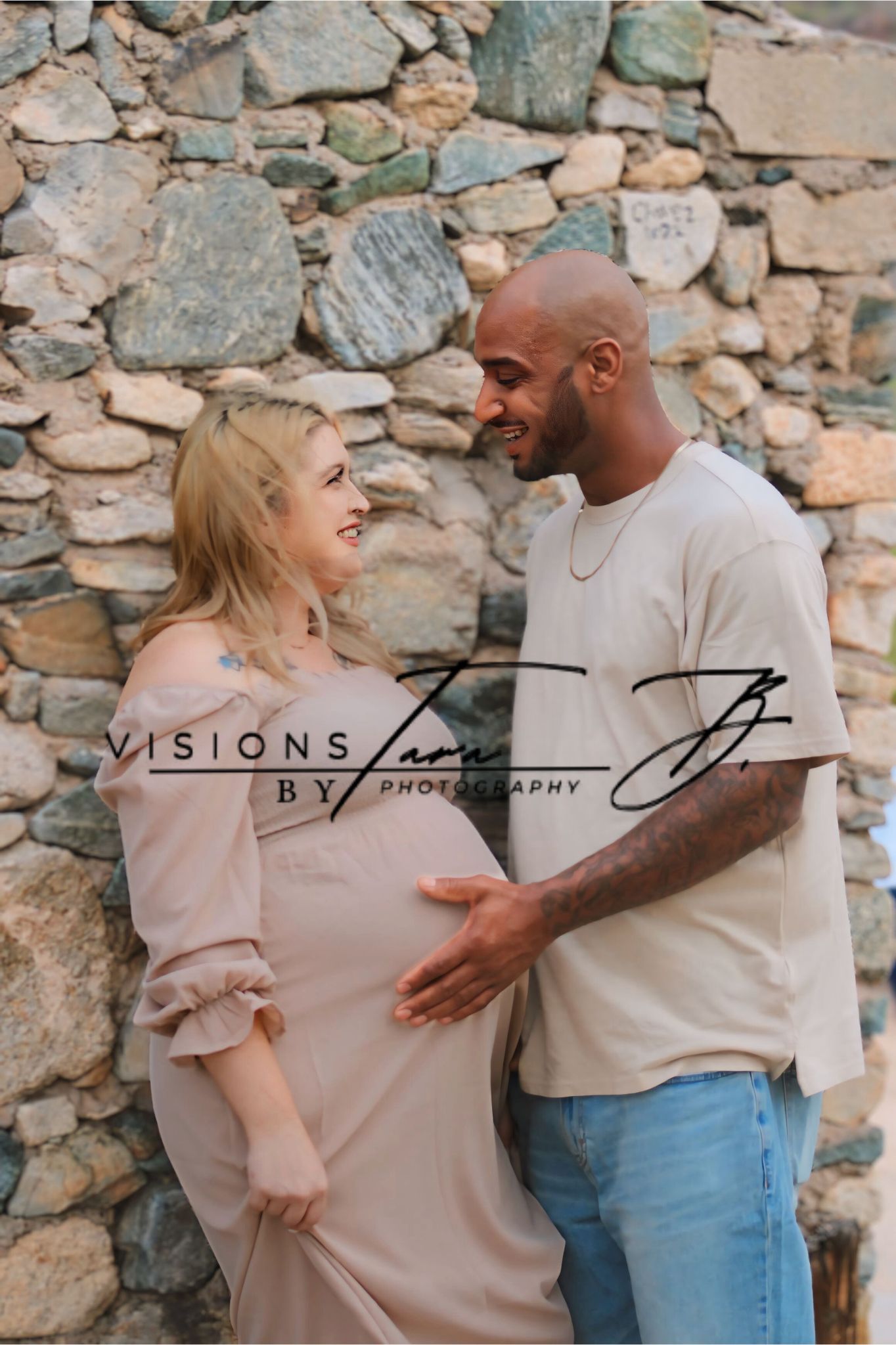 Expecting couple laughing together as they hold the baby bump, sharing a sweet and natural maternity moment by a stone wall.