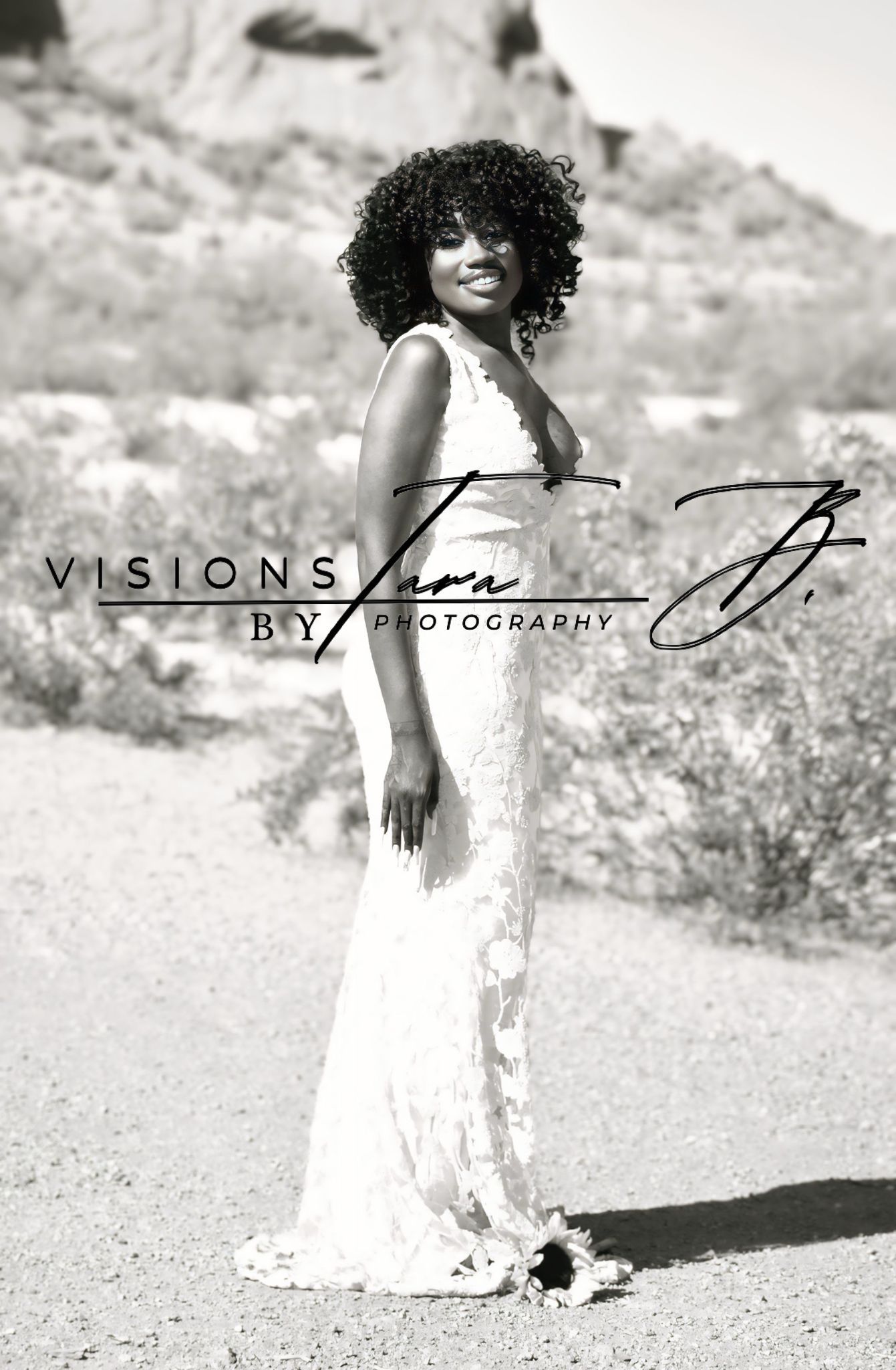 Black-and-white portrait of a woman in a gown standing in the desert, glowing in bright sunlight with a soft, serene pose.