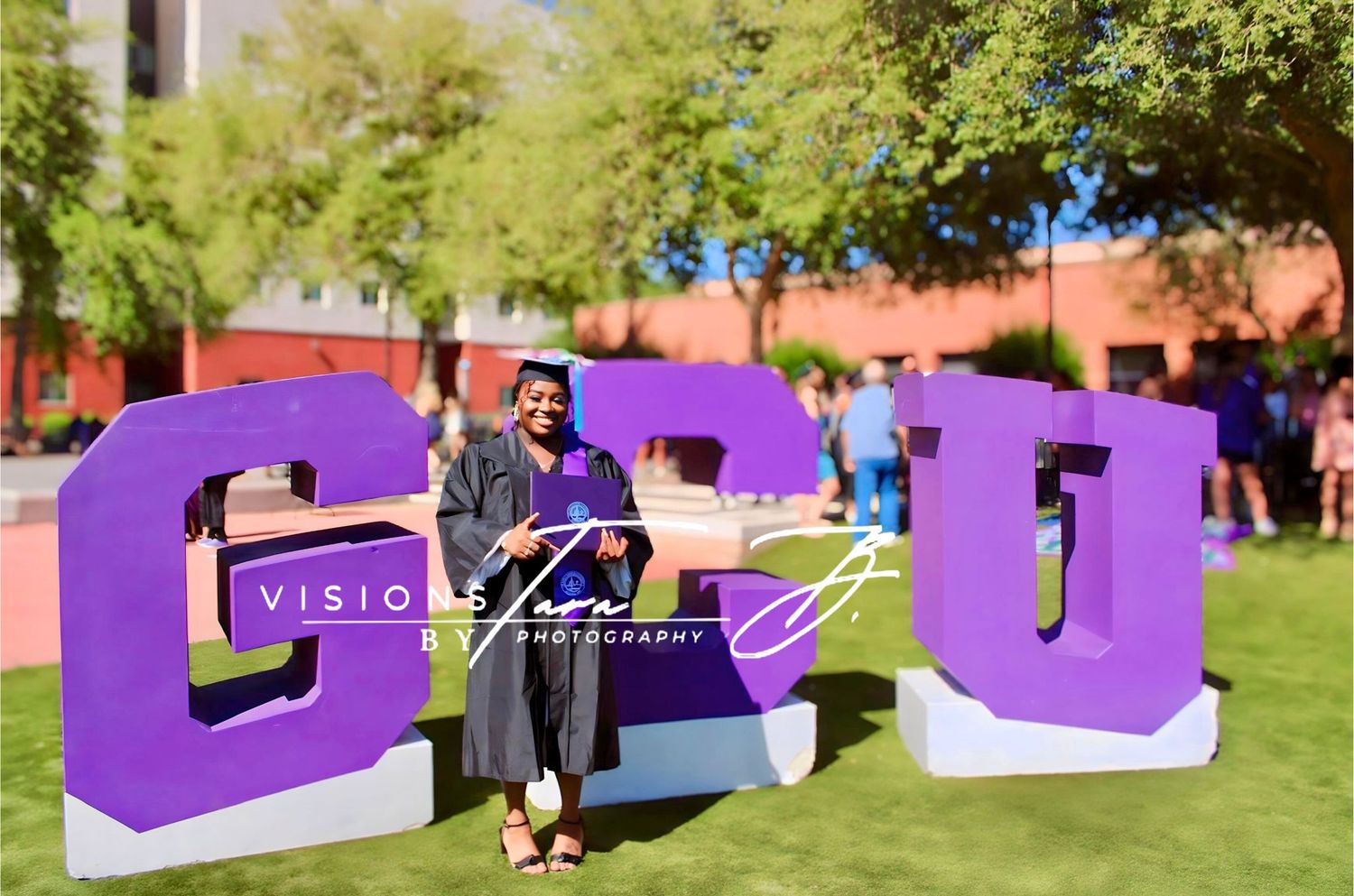 Graduate standing in front of large purple GCU letters on a sunny campus lawn, celebrating their achievement.