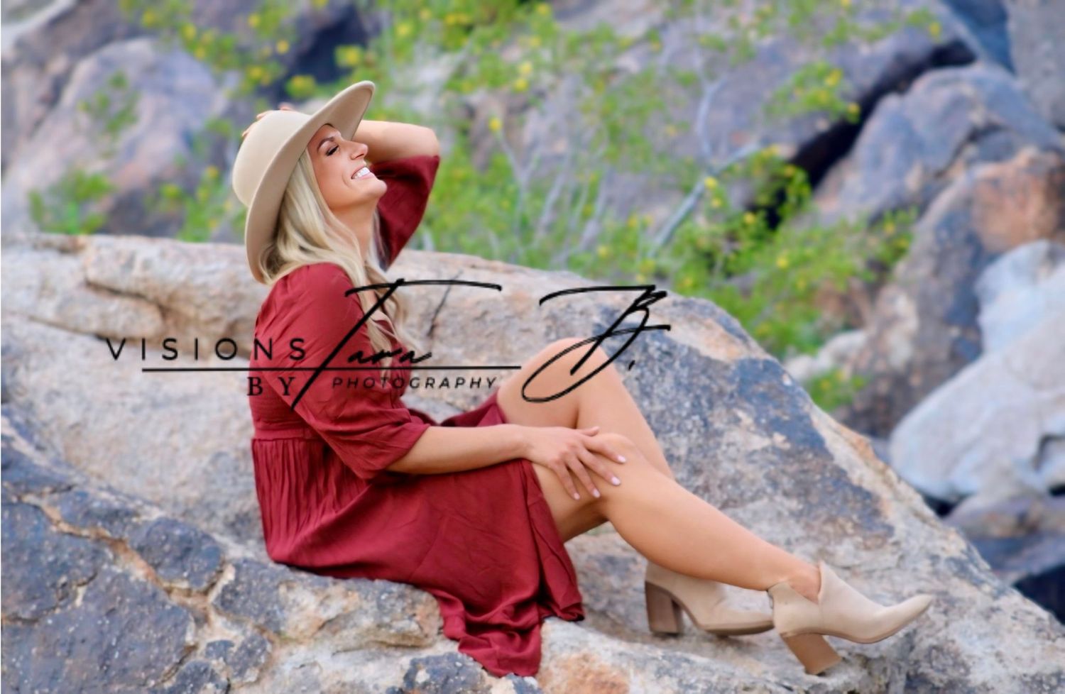Woman in a flowing red dress sitting on sunlit rocks, posing confidently with her hat held against desert scenery.